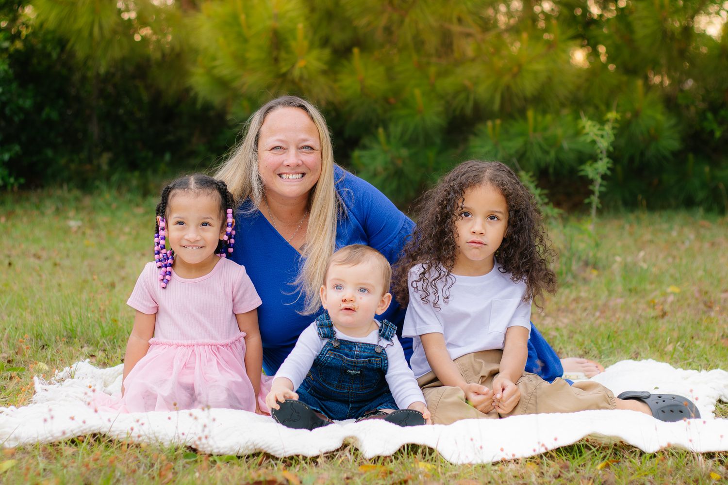 Smiling blonde woman poses with three young multiracial children on blanket in sunny outdoor park.