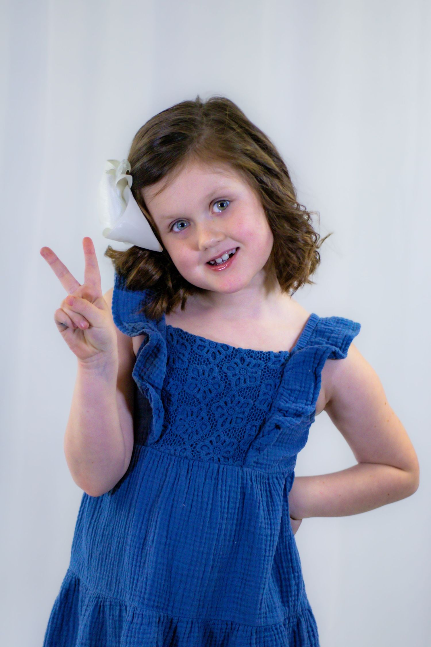 Cheerful girl in blue dress flashing peace sign with white bow in curly hair.