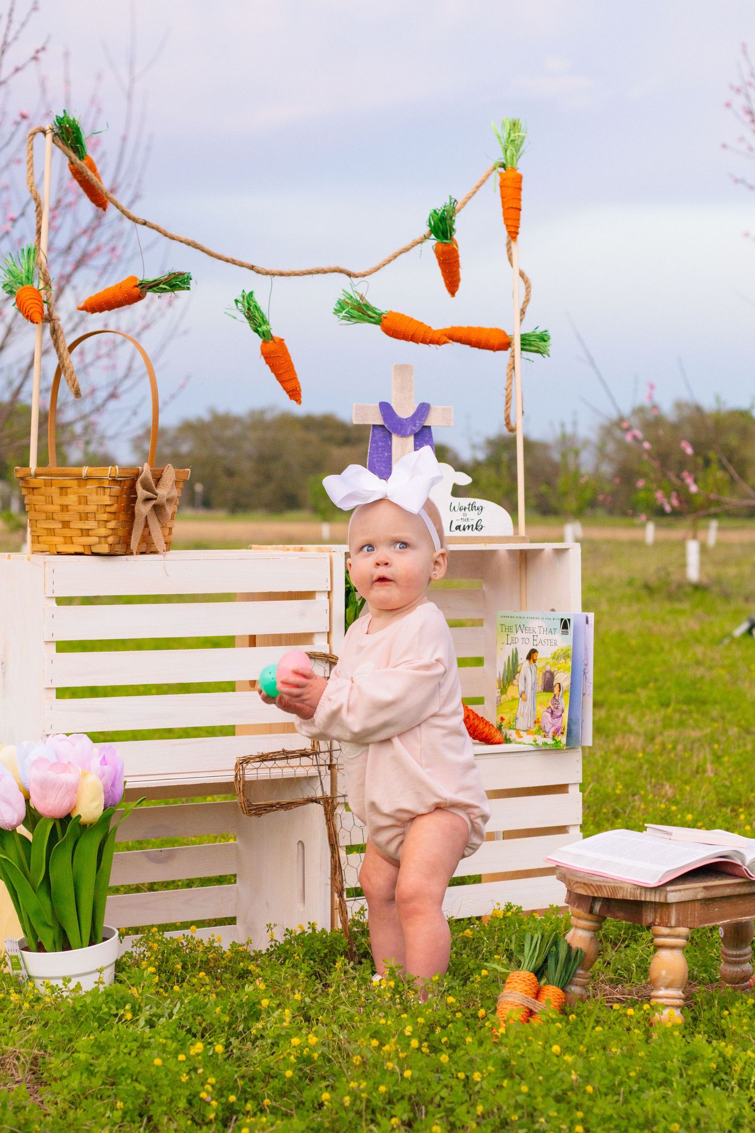 Baby in pink romper with white bow stands near white crate with carrot garland decor.