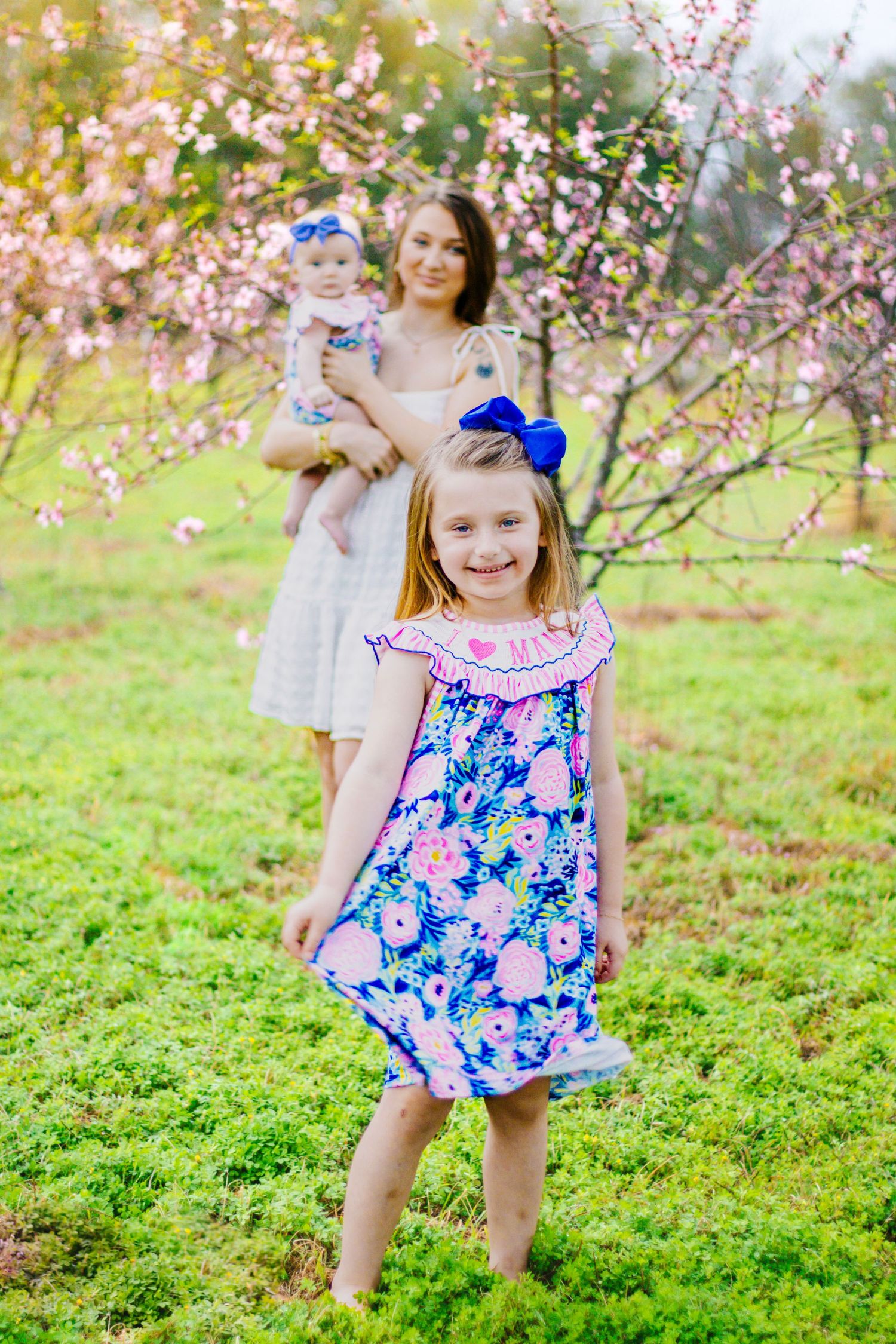 Young girl in blue floral dress smiling as mother holds baby among pink cherry blossom trees outdoors.
