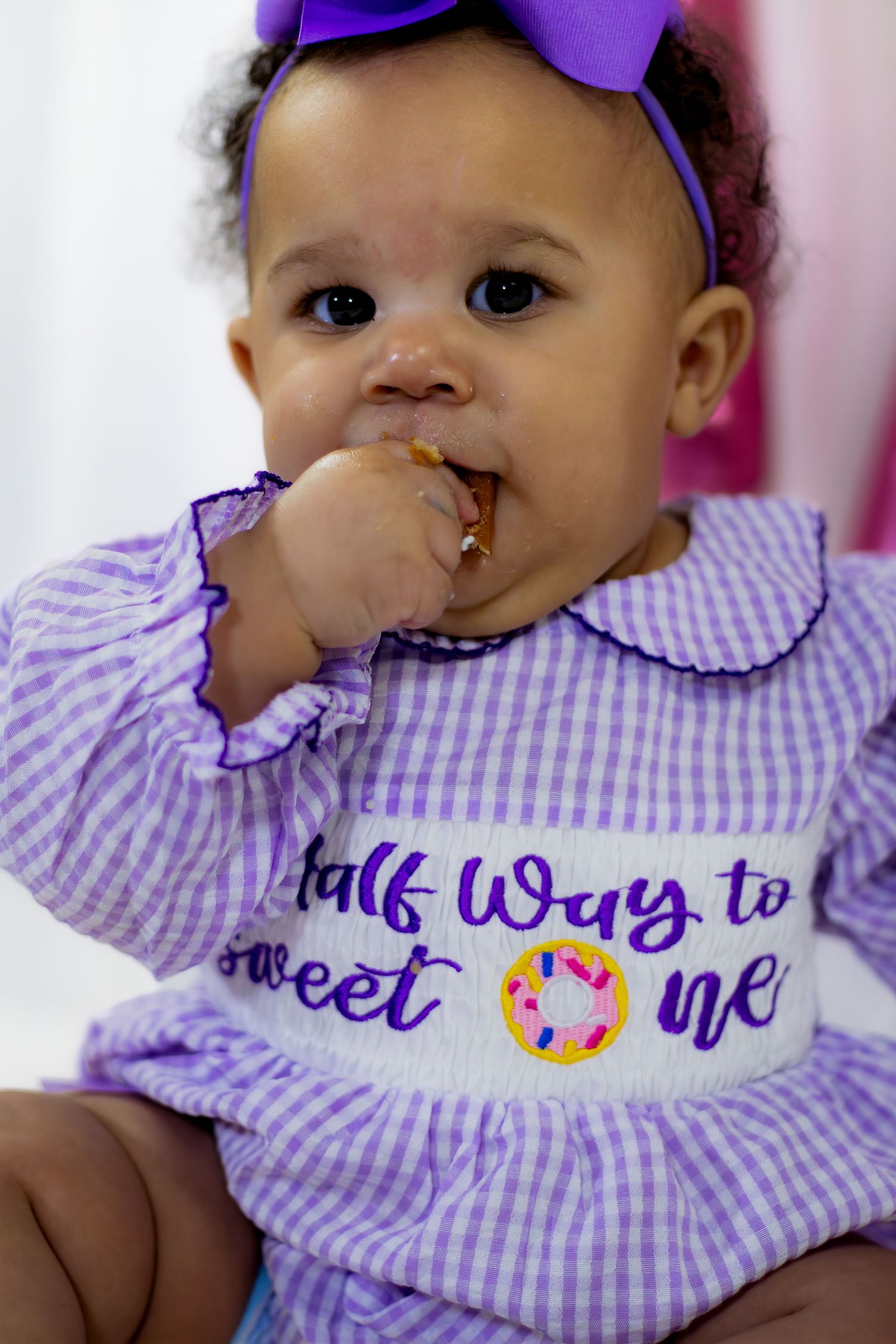 Close-up portrait of baby in Half Way to Sweet One purple gingham romper eating donut.