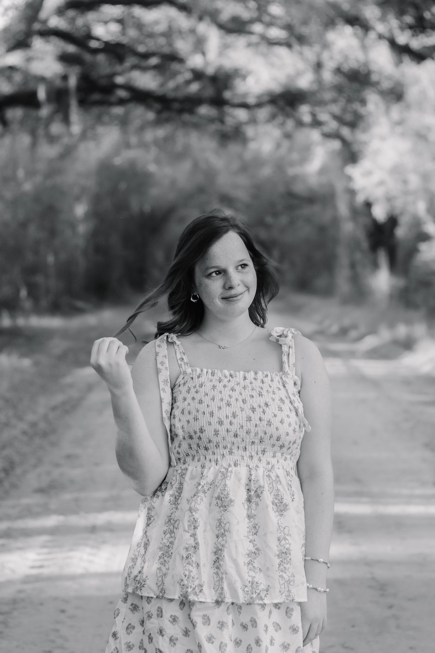 Black and white portrait of a smiling girl in a floral dress on a shaded dirt road.