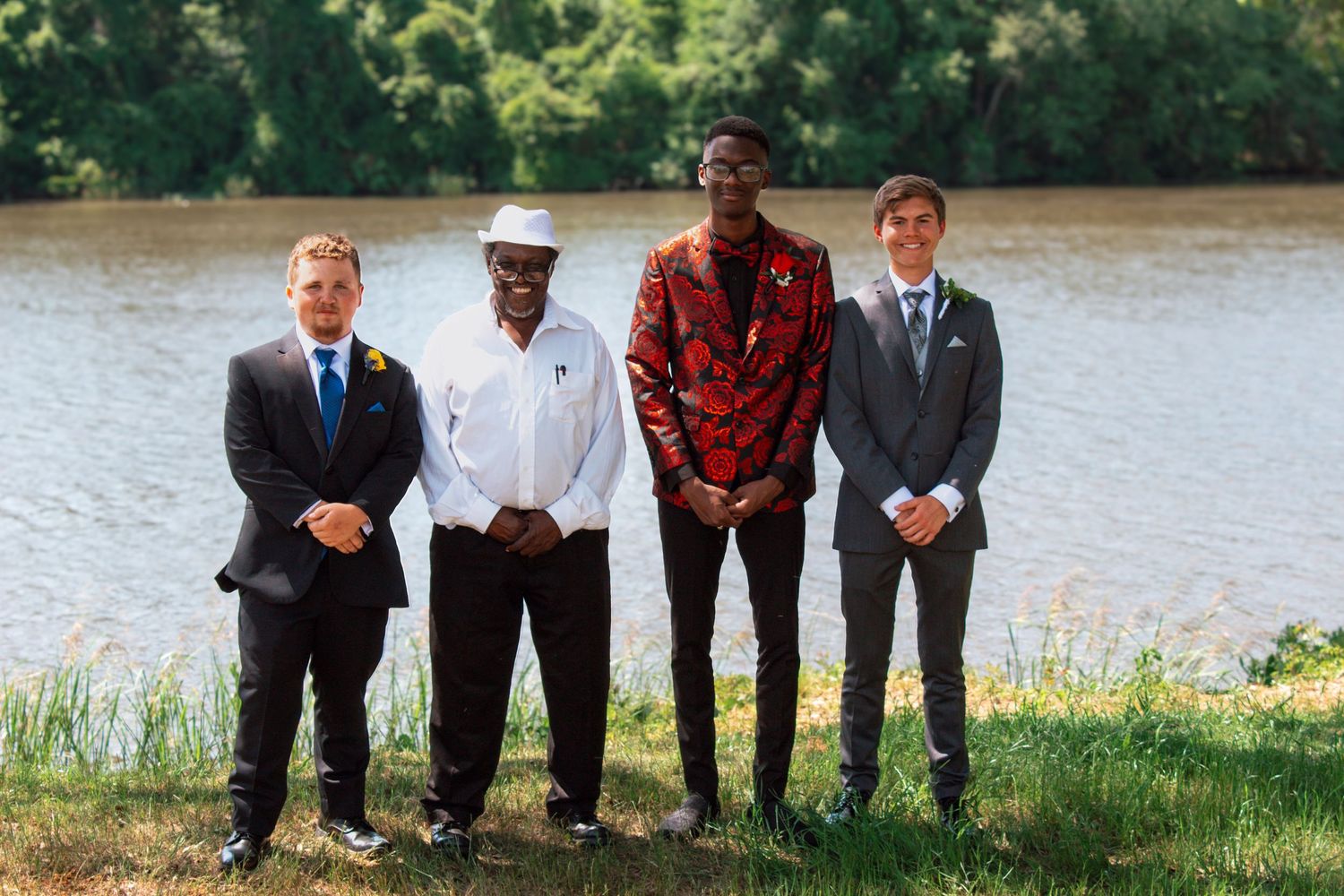 Four males pose near a river, three in formal prom attire and one in casual white outfit.
