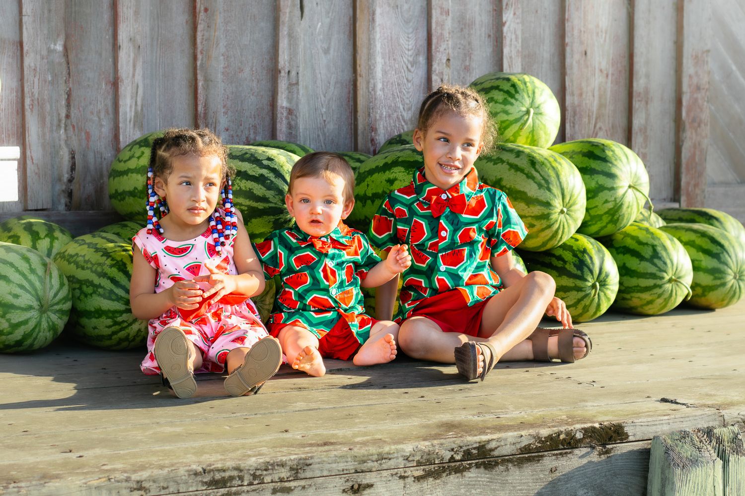Three young children in watermelon print outfits sit together on wooden deck with pile of watermelons behind them.