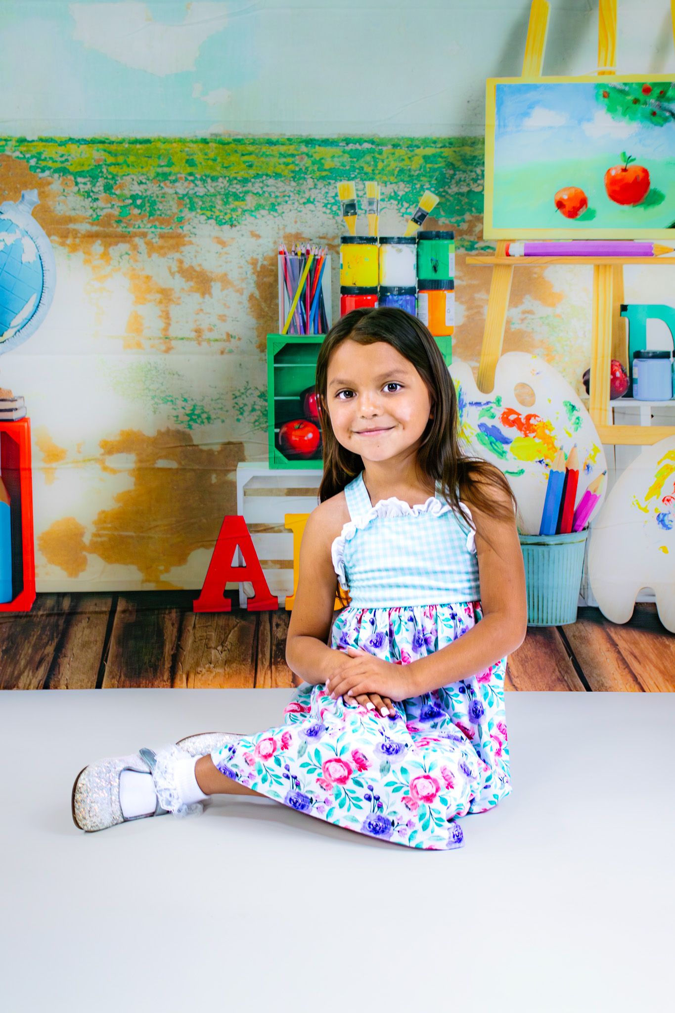 Young girl in floral dress sitting on floor in colorful art classroom with paintings and school supplies.