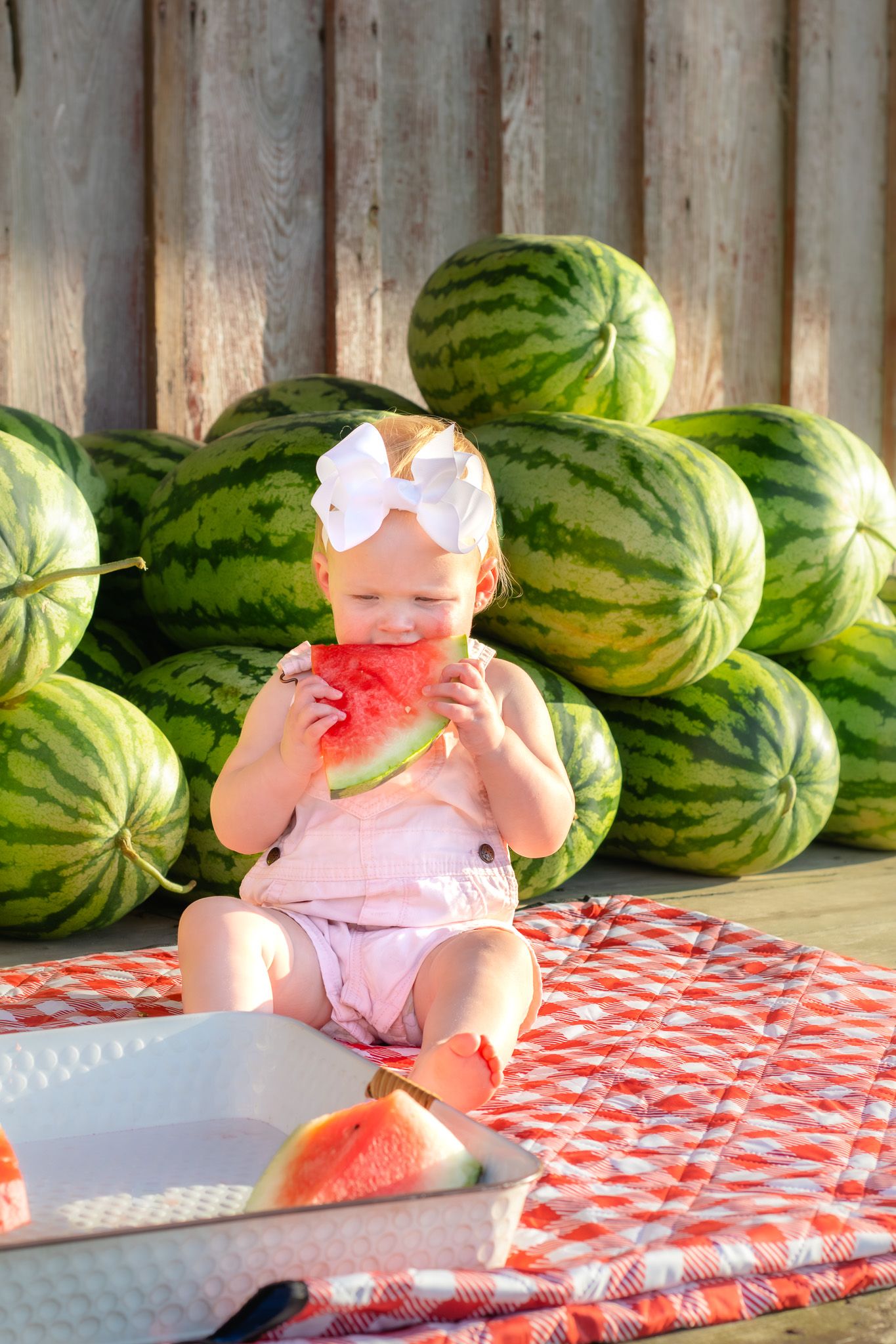 Adorable baby girl with white bow headband biting into a fresh watermelon slice outdoors near stacked watermelons.