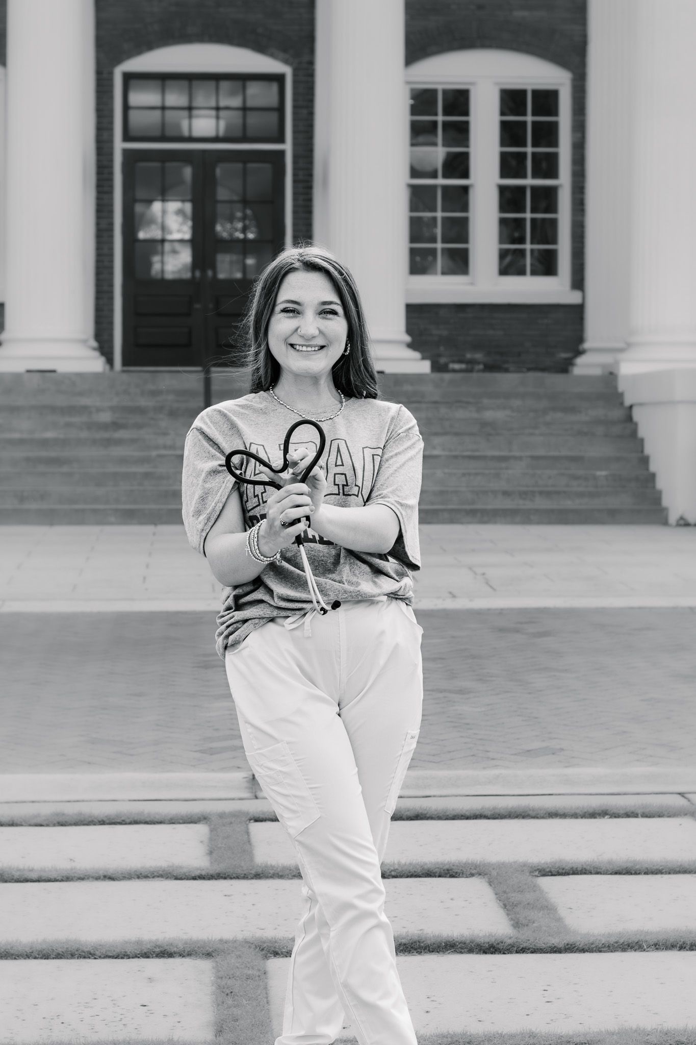 Smiling young woman in college sweatshirt and white pants holding glasses outside brick building.