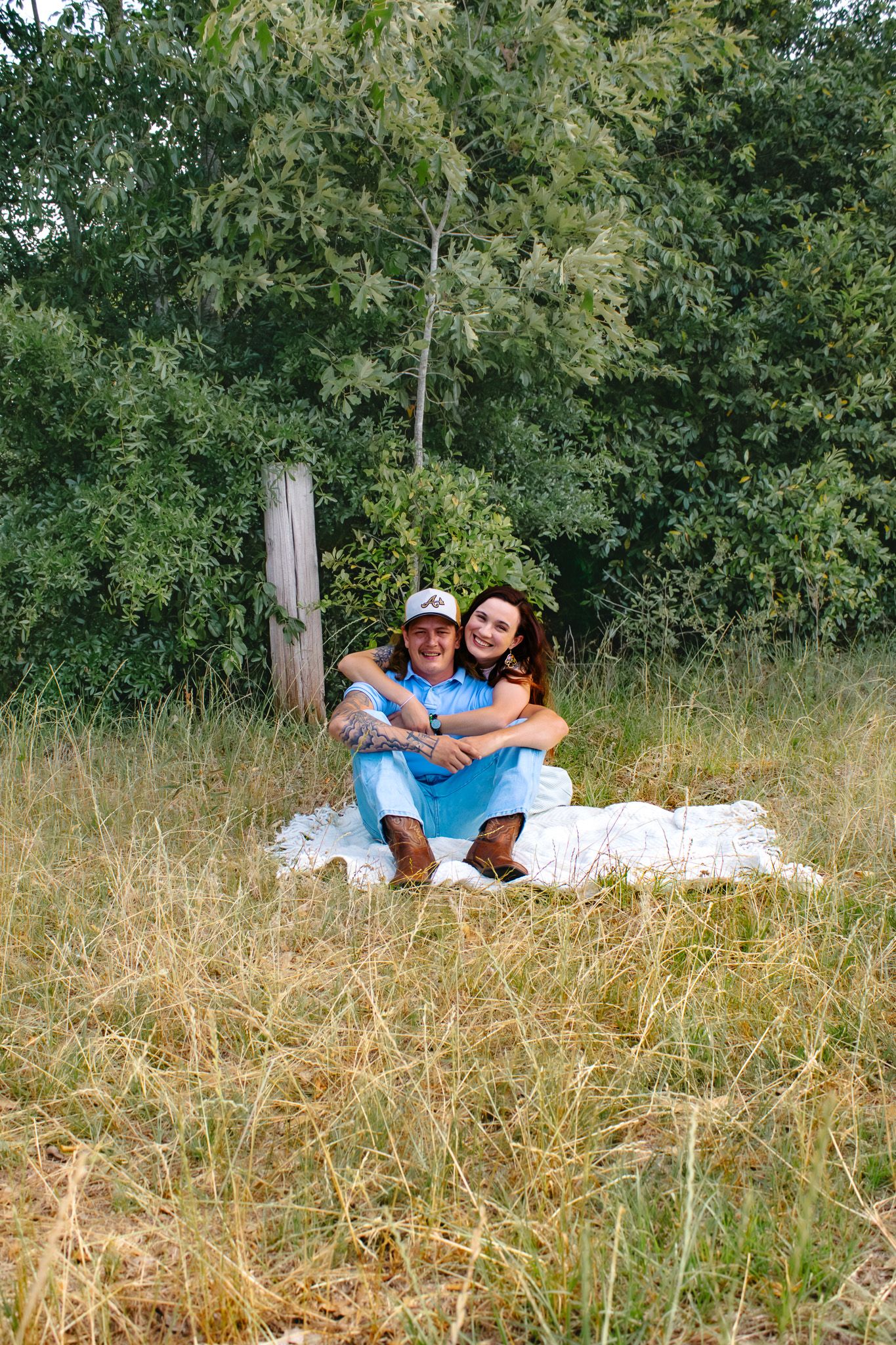 A smiling couple sits together on a white blanket in a golden grassy field surrounded by lush green trees.