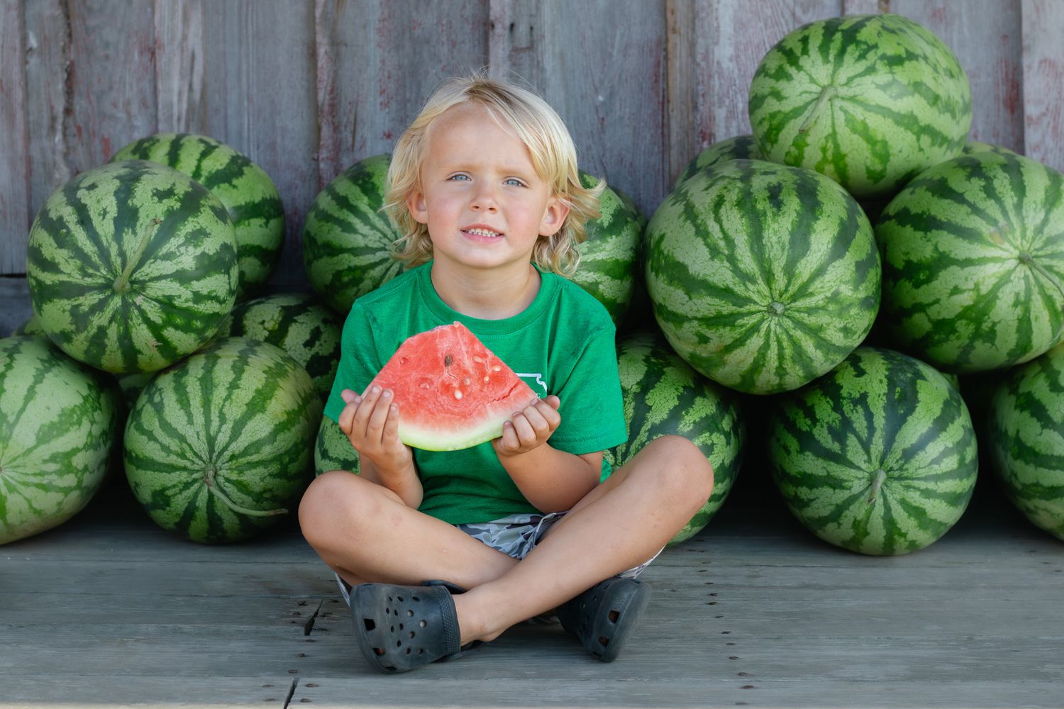 Smiling blonde boy in green shirt sitting cross-legged holding watermelon slice surrounded by pile of whole watermelons.