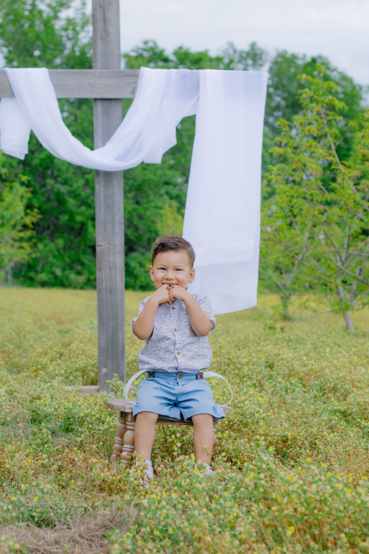 Cute toddler boy in blue shorts and suspenders sits on small stool in flower field with white fabric on wooden cross behind him.