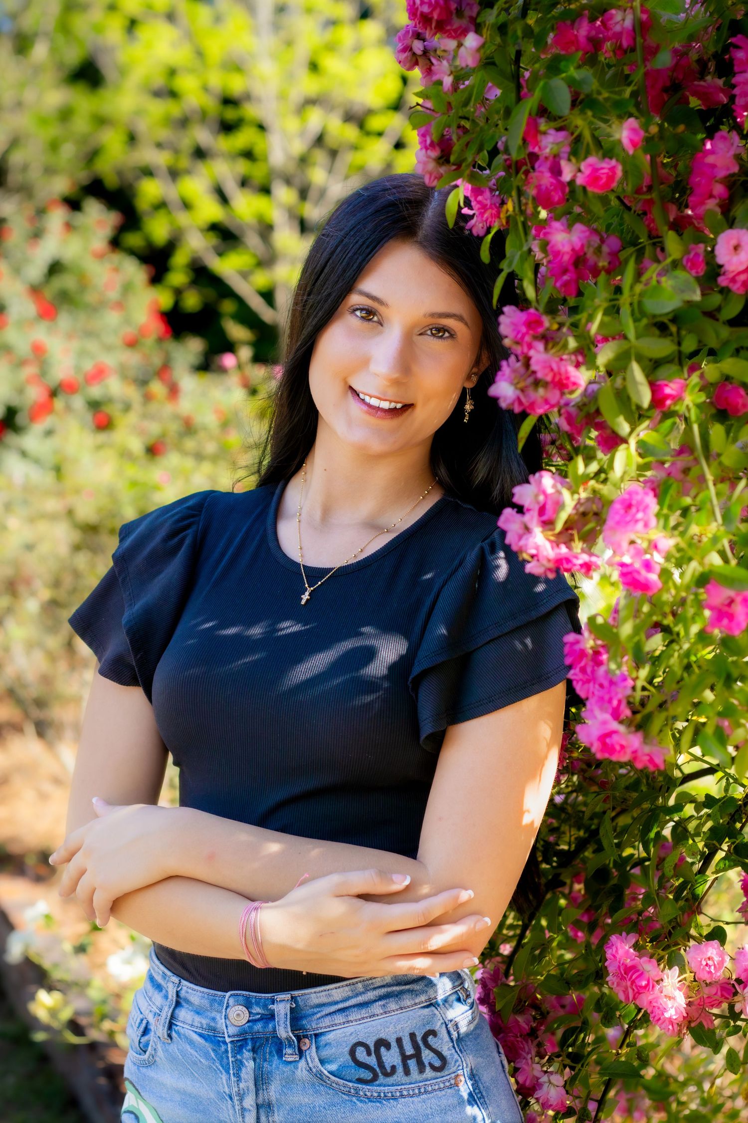 Smiling young woman in black top with SCHS jeans stands by vibrant pink flowering bushes outdoors.