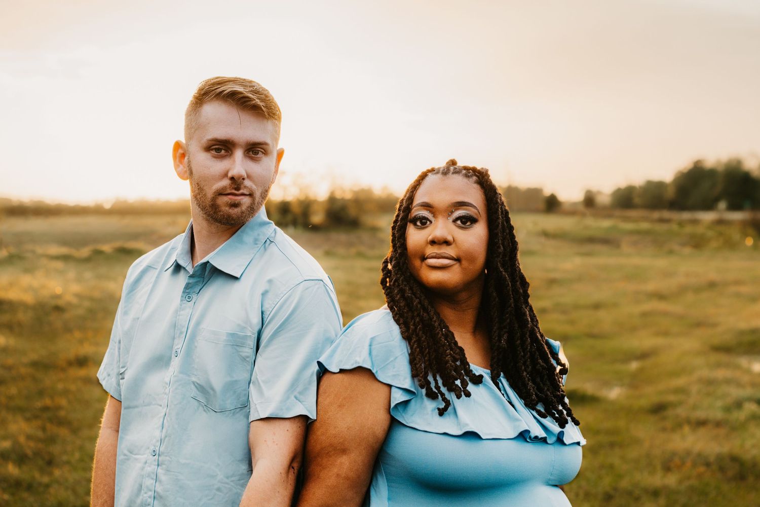 Interracial couple poses back to back in golden hour field, wearing matching light blue outfits.
