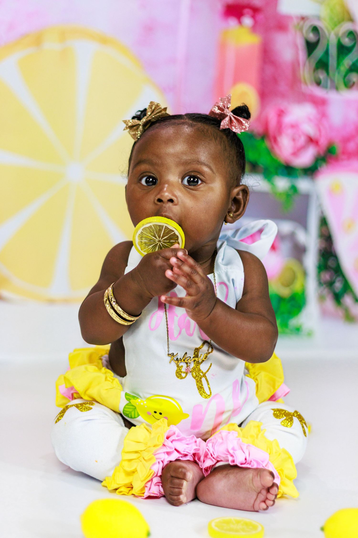 Baby girl in a lemon-print outfit sitting cross-legged and chewing on a lemon slice prop surrounded by lemons.