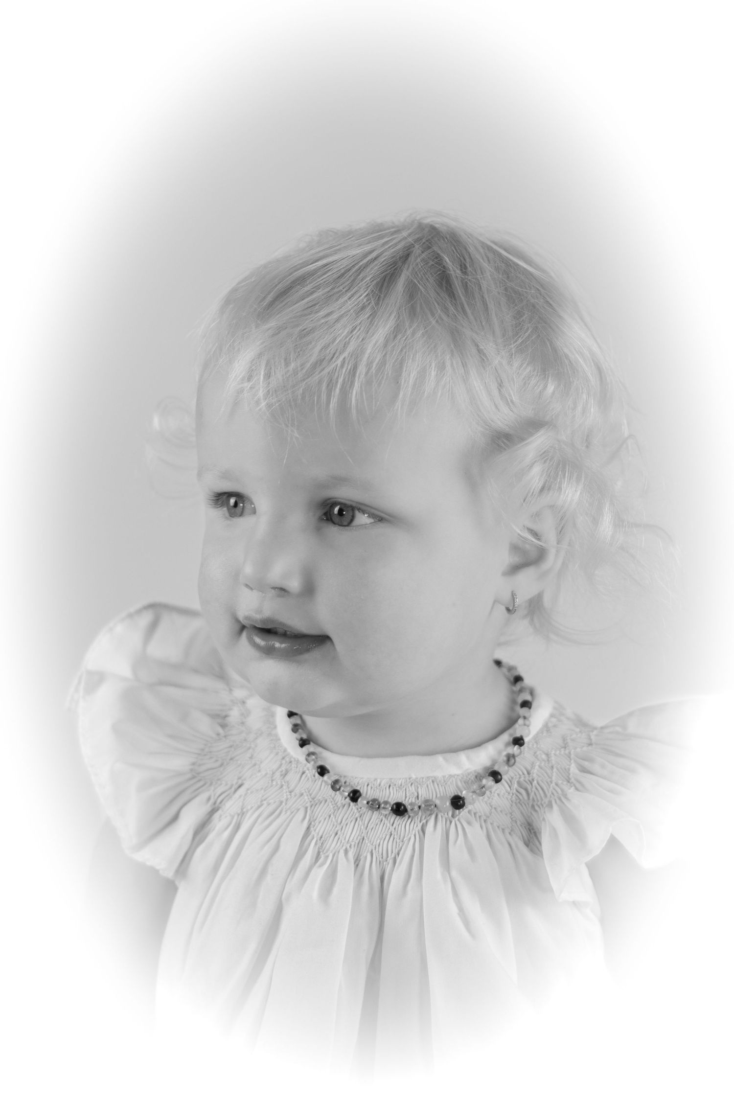 Black and white portrait of a smiling toddler girl wearing a white smocked dress and beaded necklace.