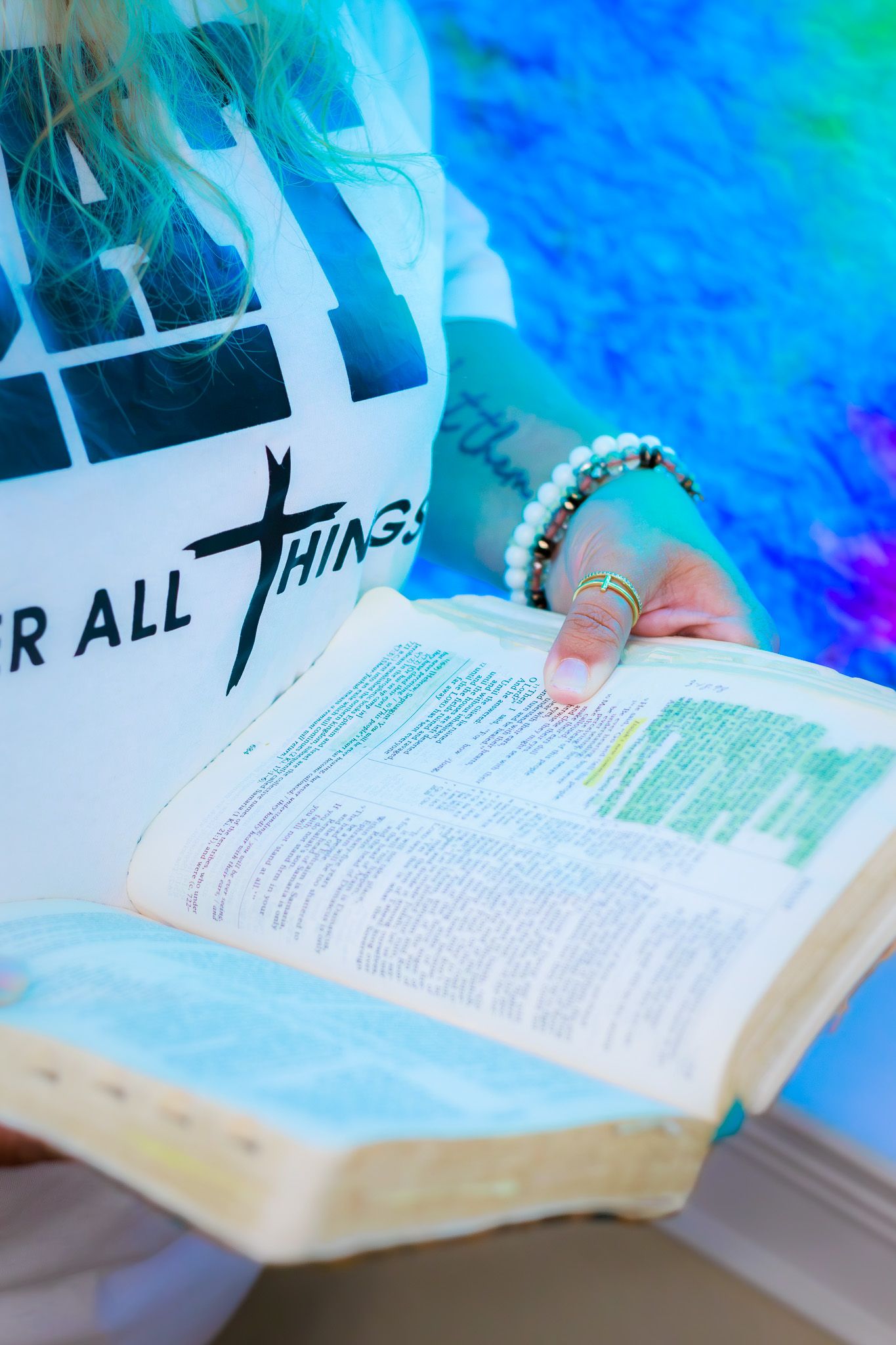 Close-up of hands holding open highlighted Bible wearing colorful bracelets near stained glass window.