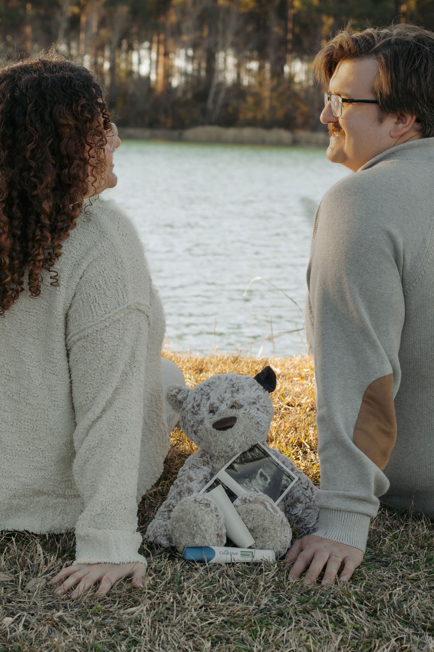 Couple sitting by lake with teddy bear and ultrasound photos between them.