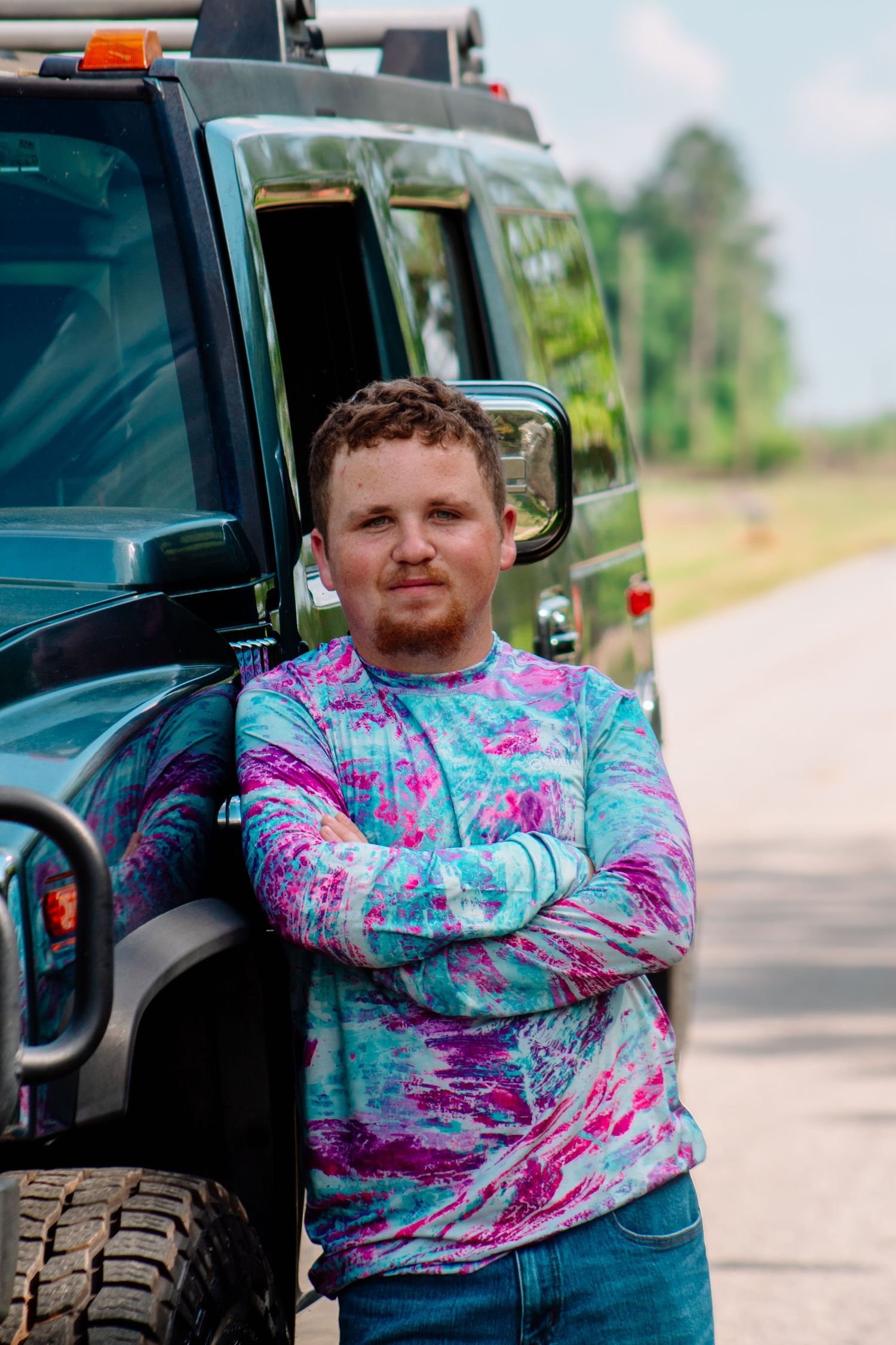 Young man in pink and blue tie-dye long sleeve shirt standing with arms crossed beside a Hummer.