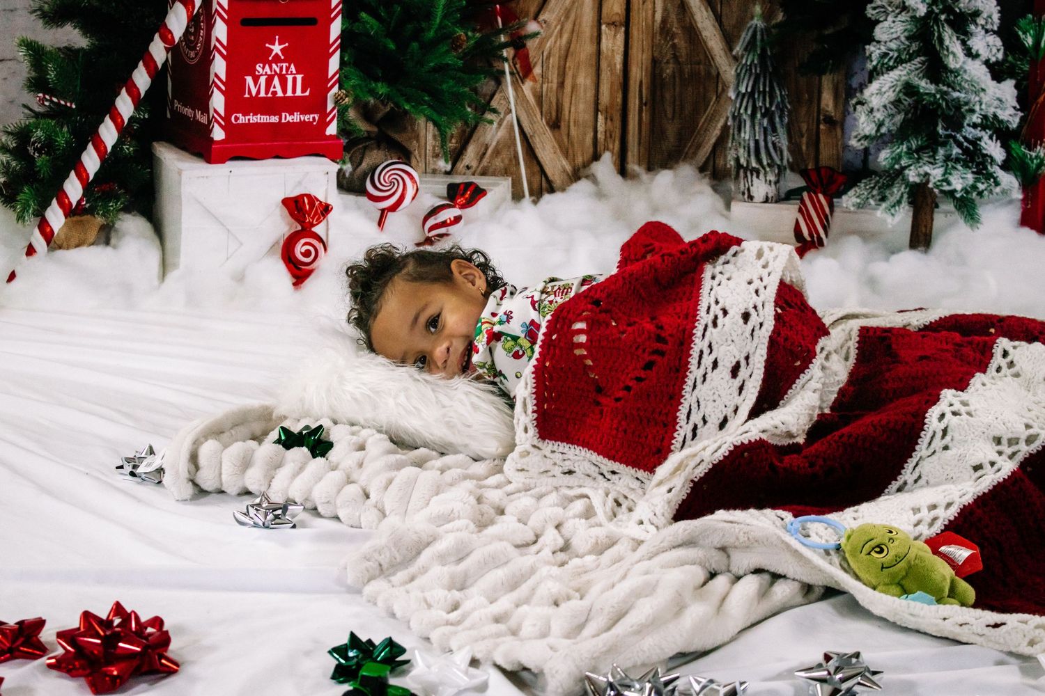 Young child wrapped in red and white blankets lying in a Christmas-themed photo studio setting.