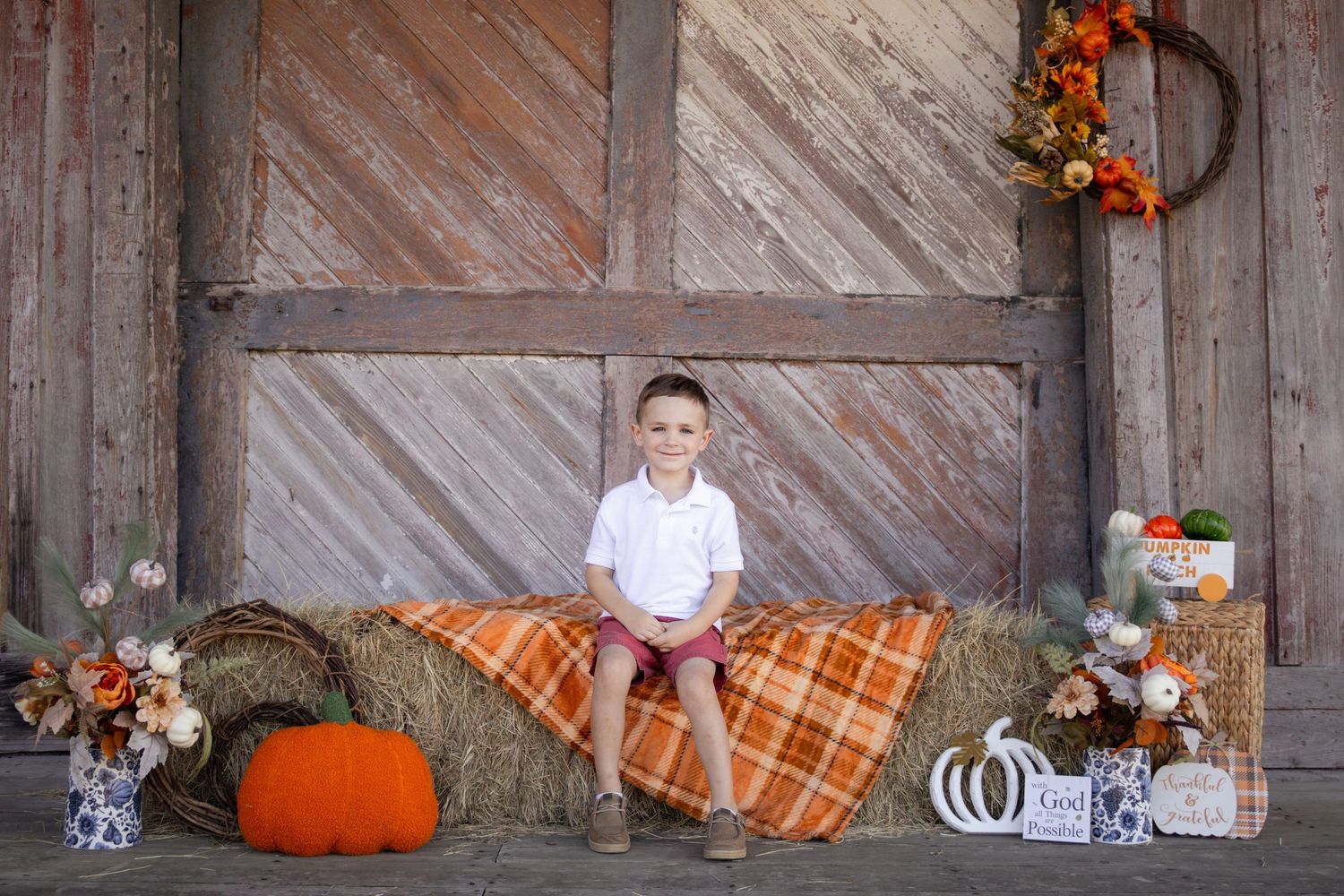 Boy in white polo sitting on hay bale with orange plaid blanket and pumpkins in fall barn photo.