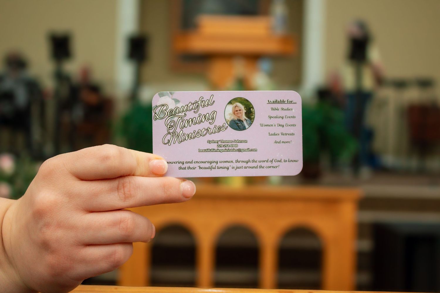 A hand holds a small decorative ministry business card in front of a church sanctuary interior.