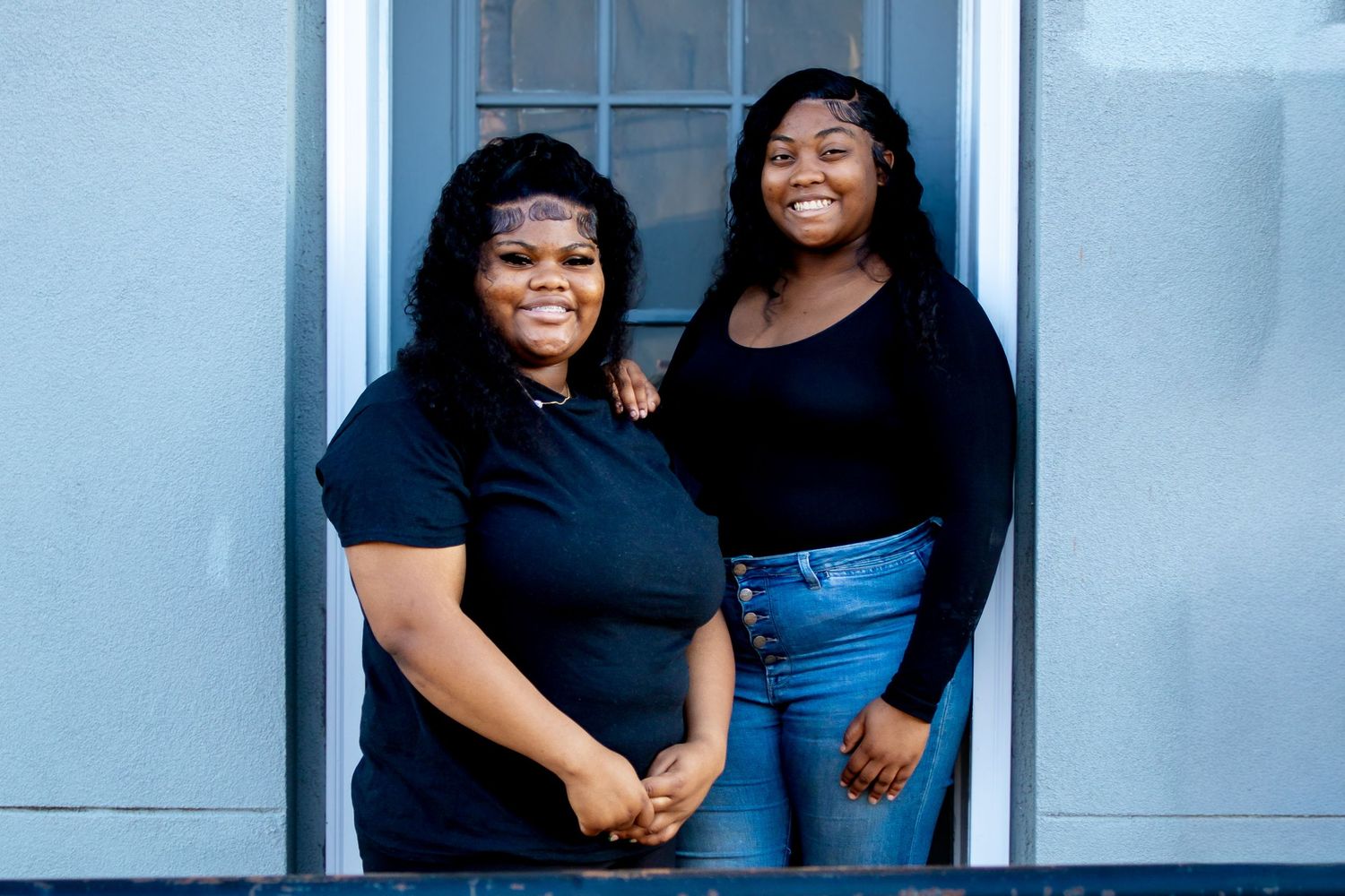 Two young Black women smiling together in matching black tops, standing in a doorway.