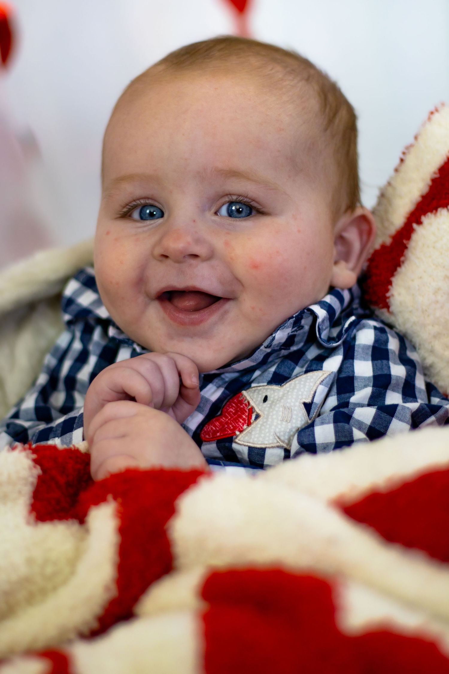 Happy blue-eyed baby laughing while wrapped in a red and white holiday blanket wearing a plaid shirt.