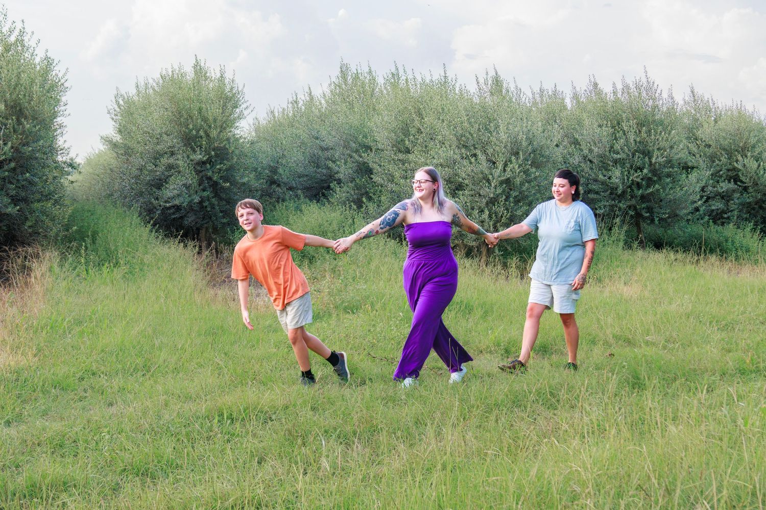 Three people holding hands and walking in a green meadow on a sunny day, woman in purple dress flanked by two men.