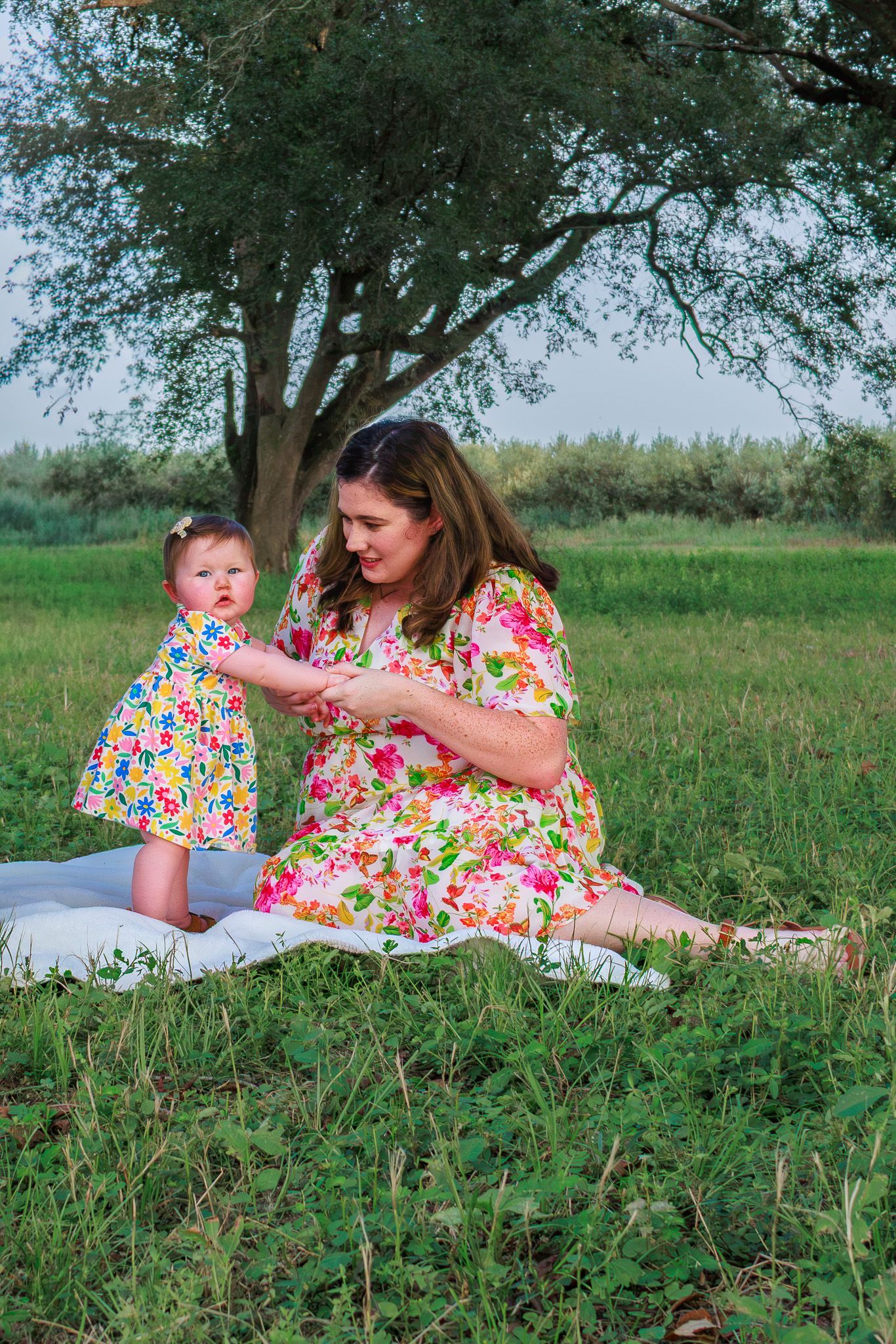Mother and baby daughter in matching floral dresses sitting on blanket in green field under large tree.