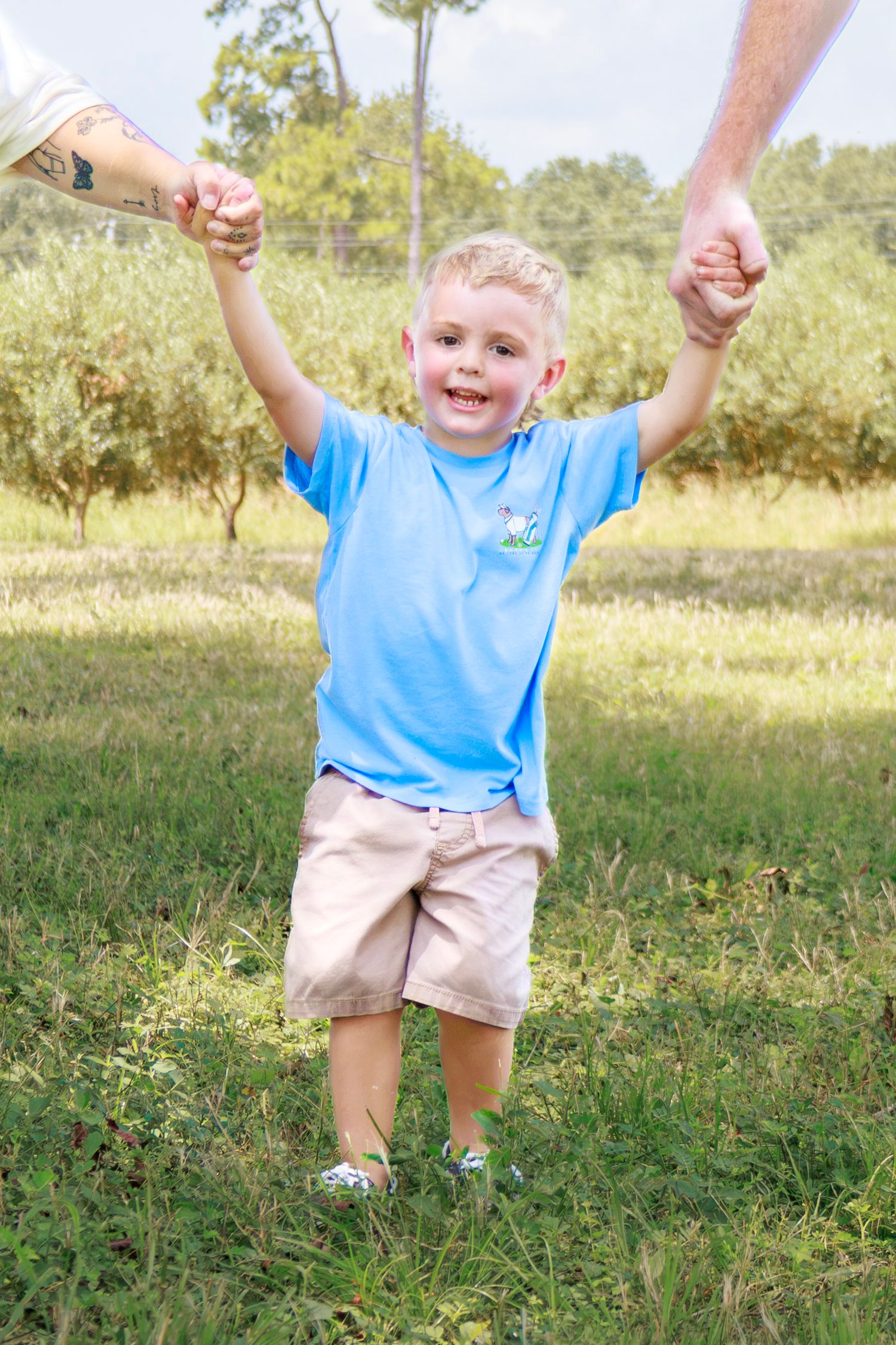 Happy blonde toddler boy in blue polo shirt holding adults hands outdoors in green park setting.
