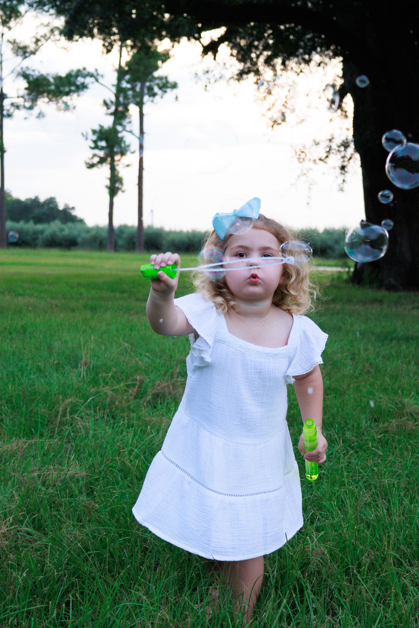Young girl in white dress blowing soap bubbles outdoors in a lush green park setting.