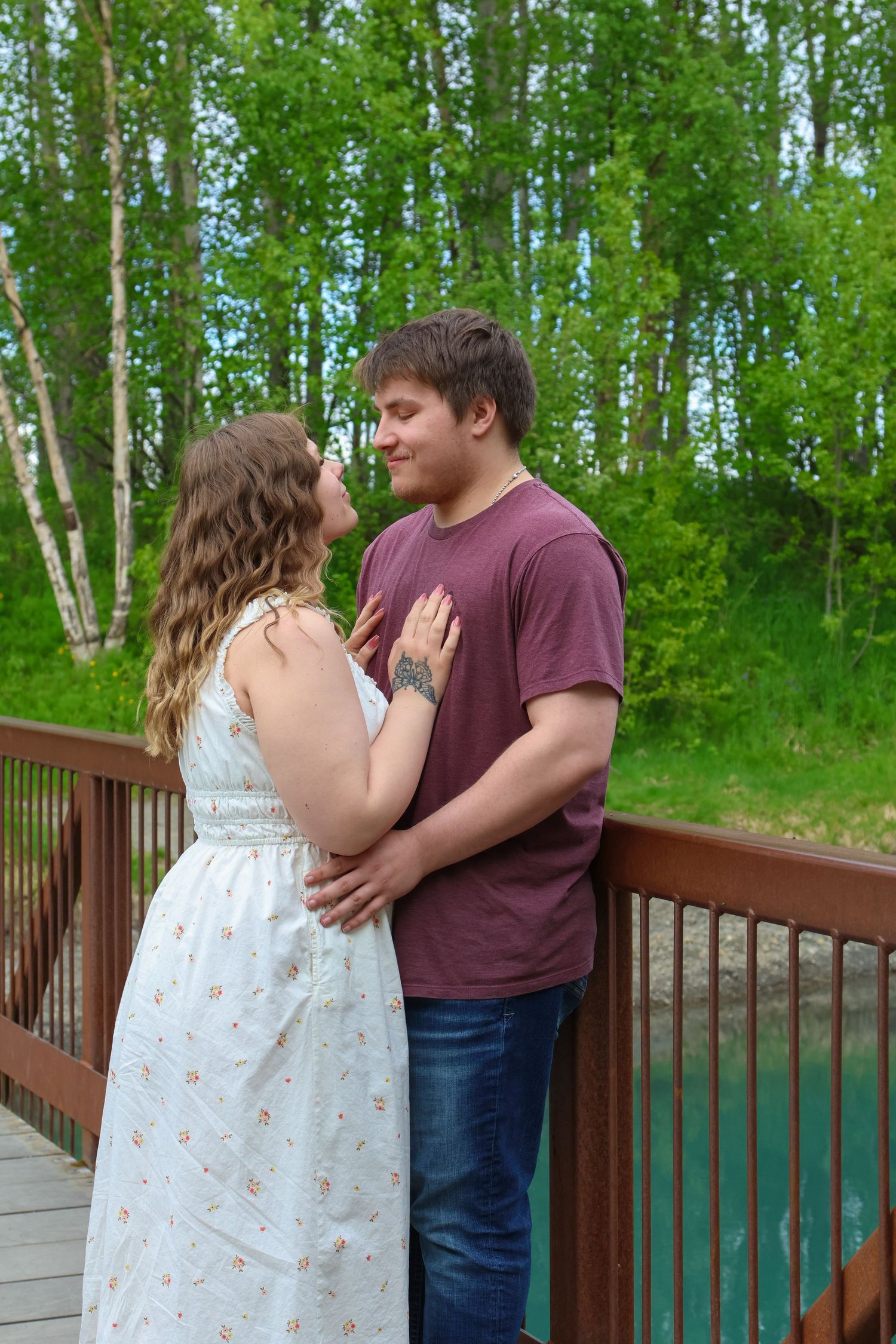 A couple shares a tender moment on a wooden deck surrounded by green birch trees in summer.