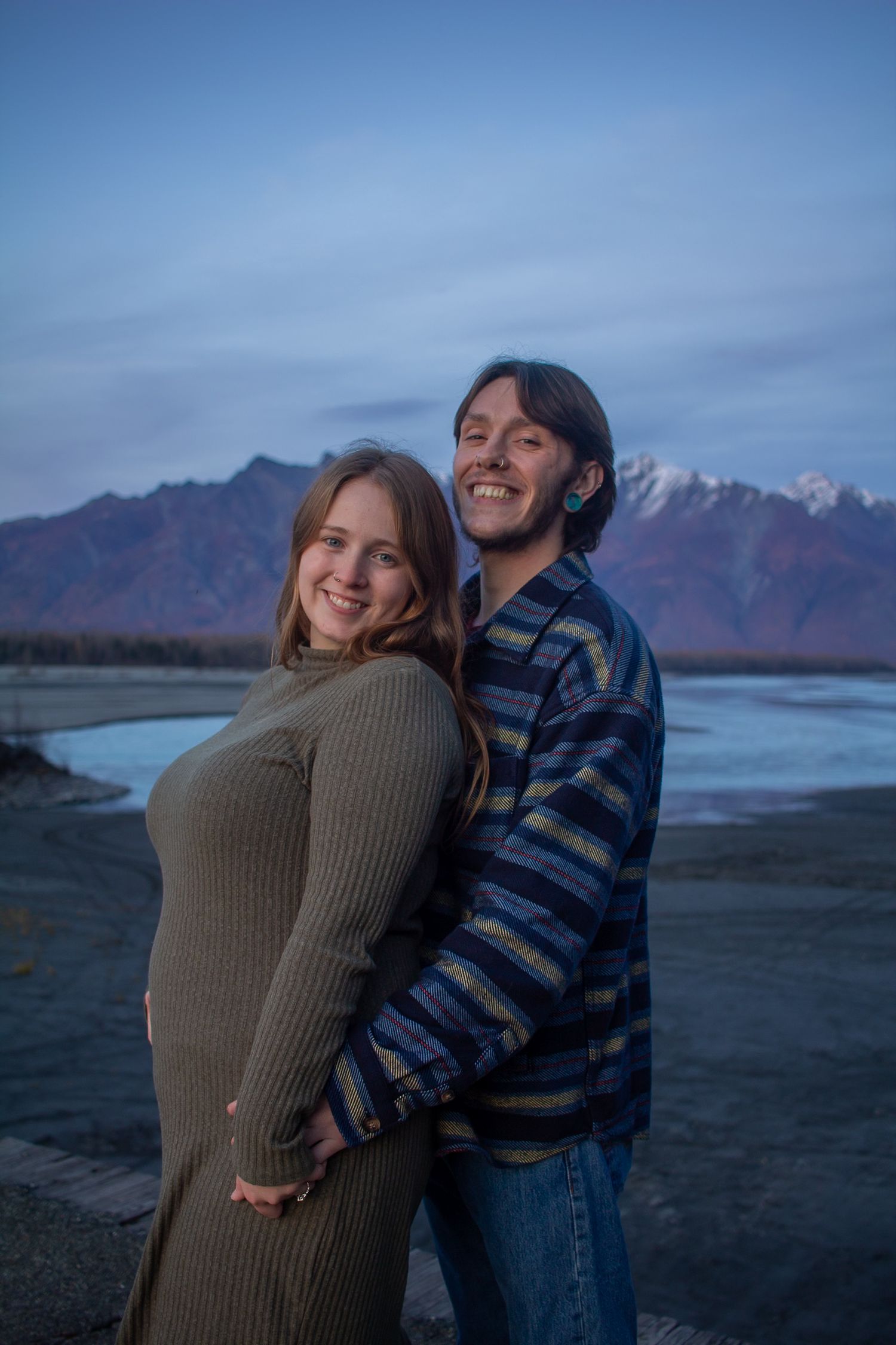 A couple stands together at dusk with snow-capped mountains and a river in the background in Alaska.