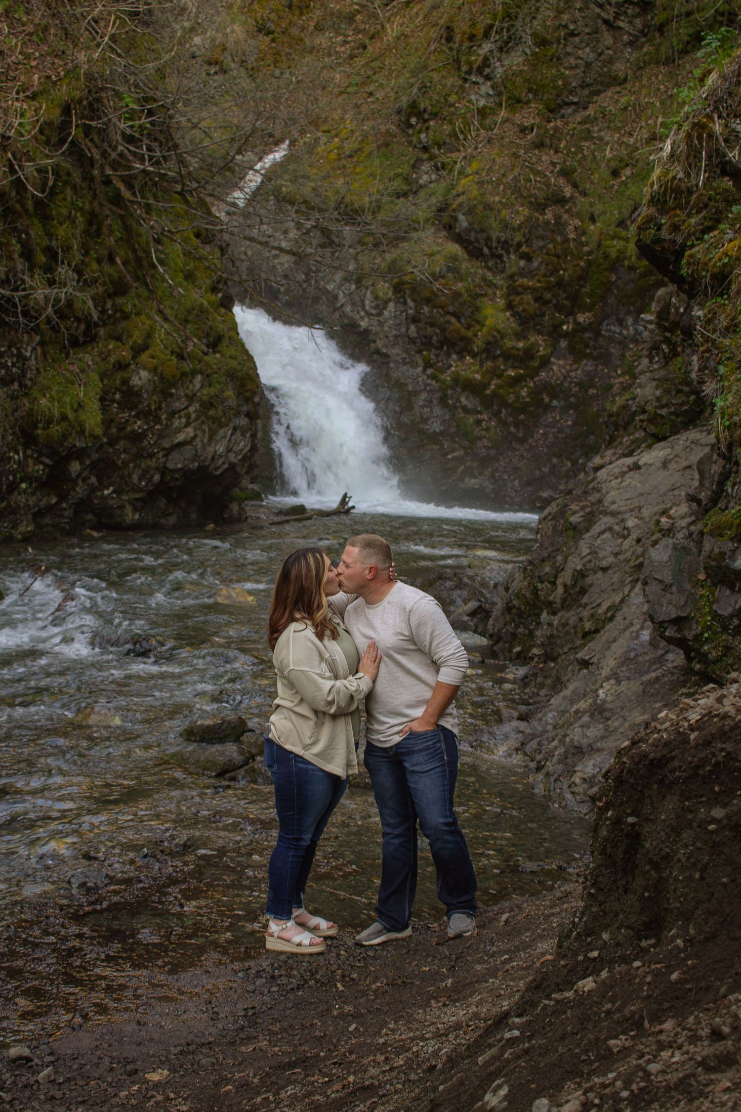Two people share an intimate moment near a cascading waterfall in a scenic forest setting.