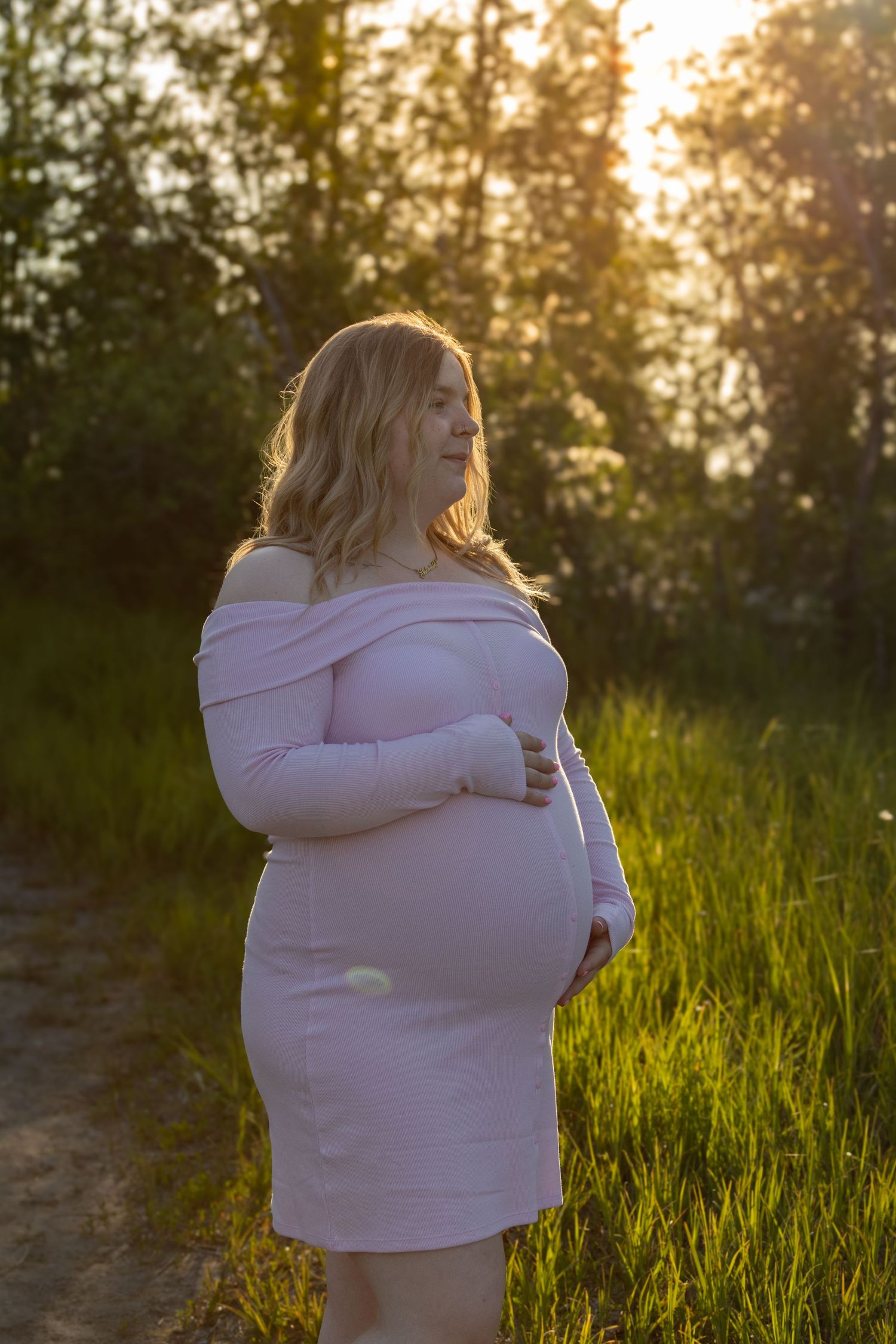 Two people share a tender embrace in a sunlit field at golden hour with tall grass and trees in the background.