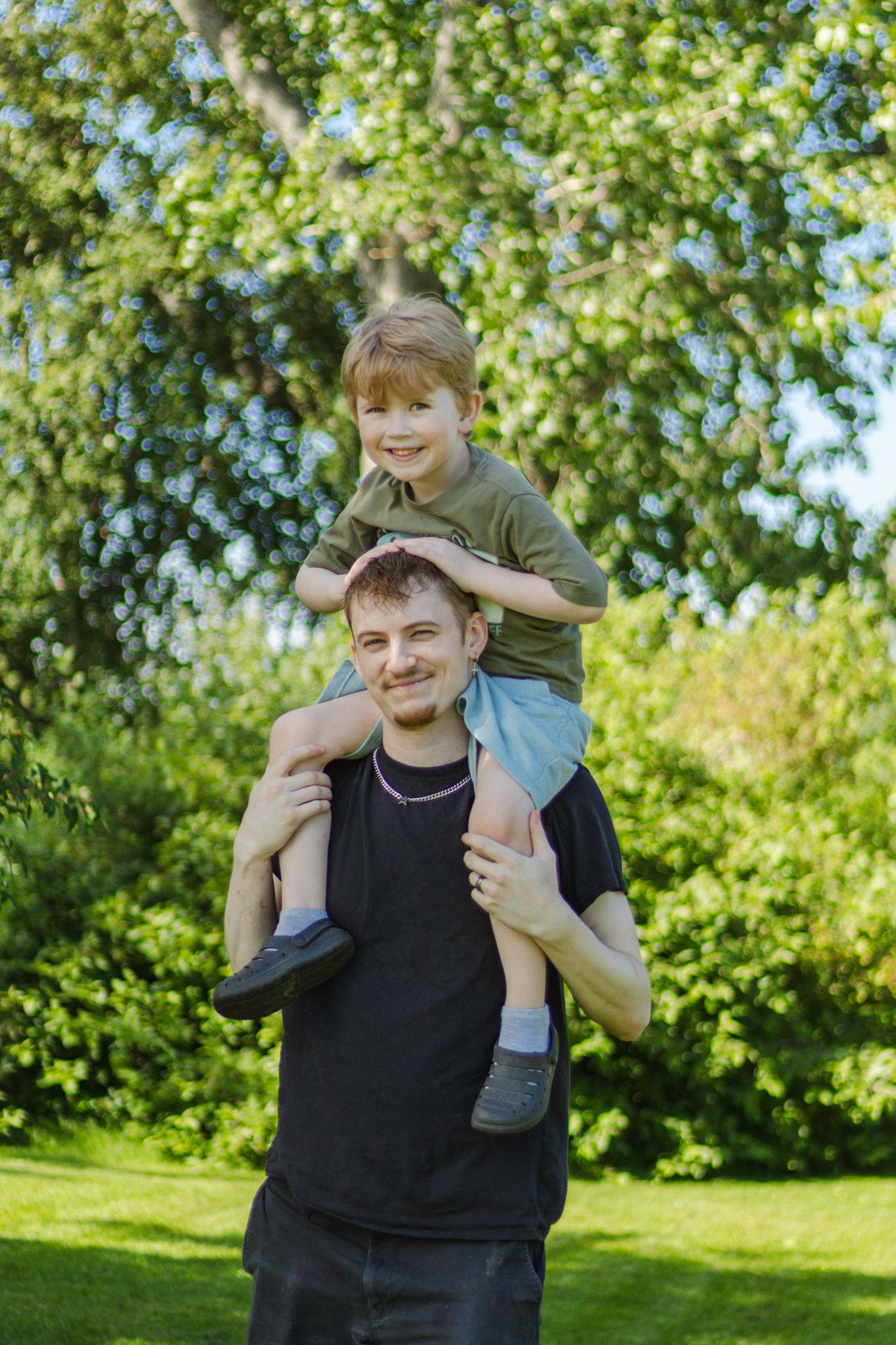 Playful outdoor scene showing piggyback ride in a sunny garden with green trees and foliage in the background.