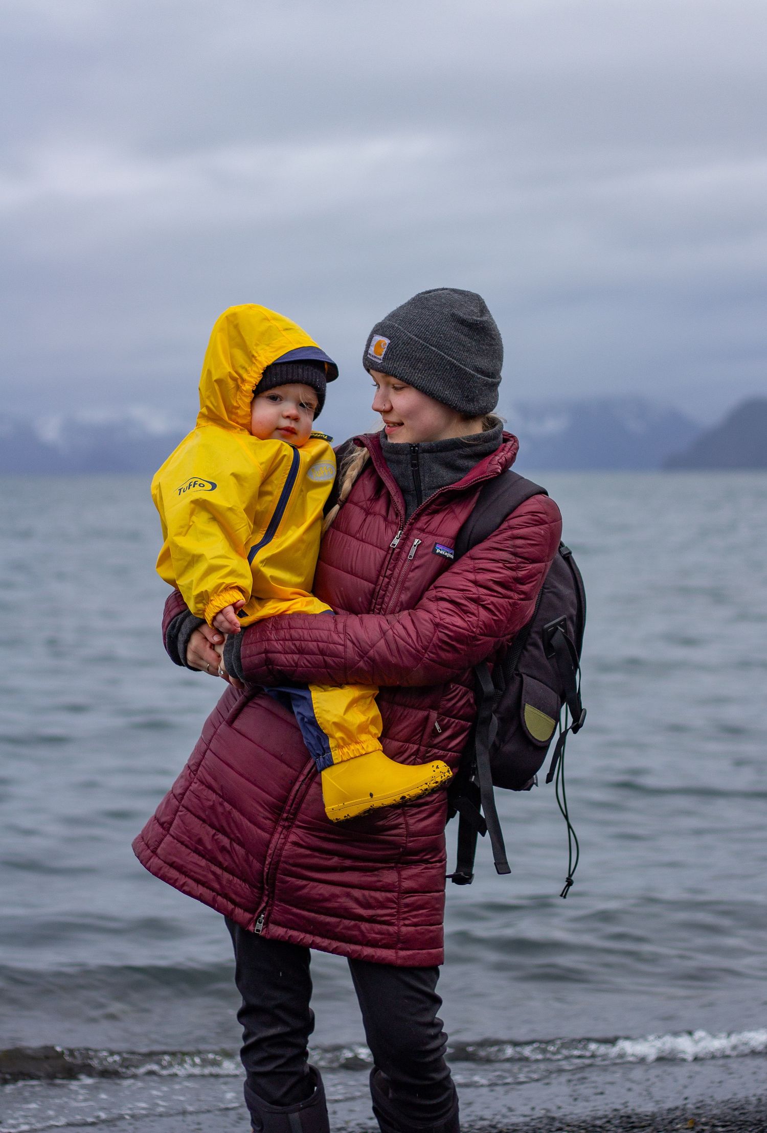 Two people in winter coats embrace on a cloudy beach with waves and mountains in the distance.