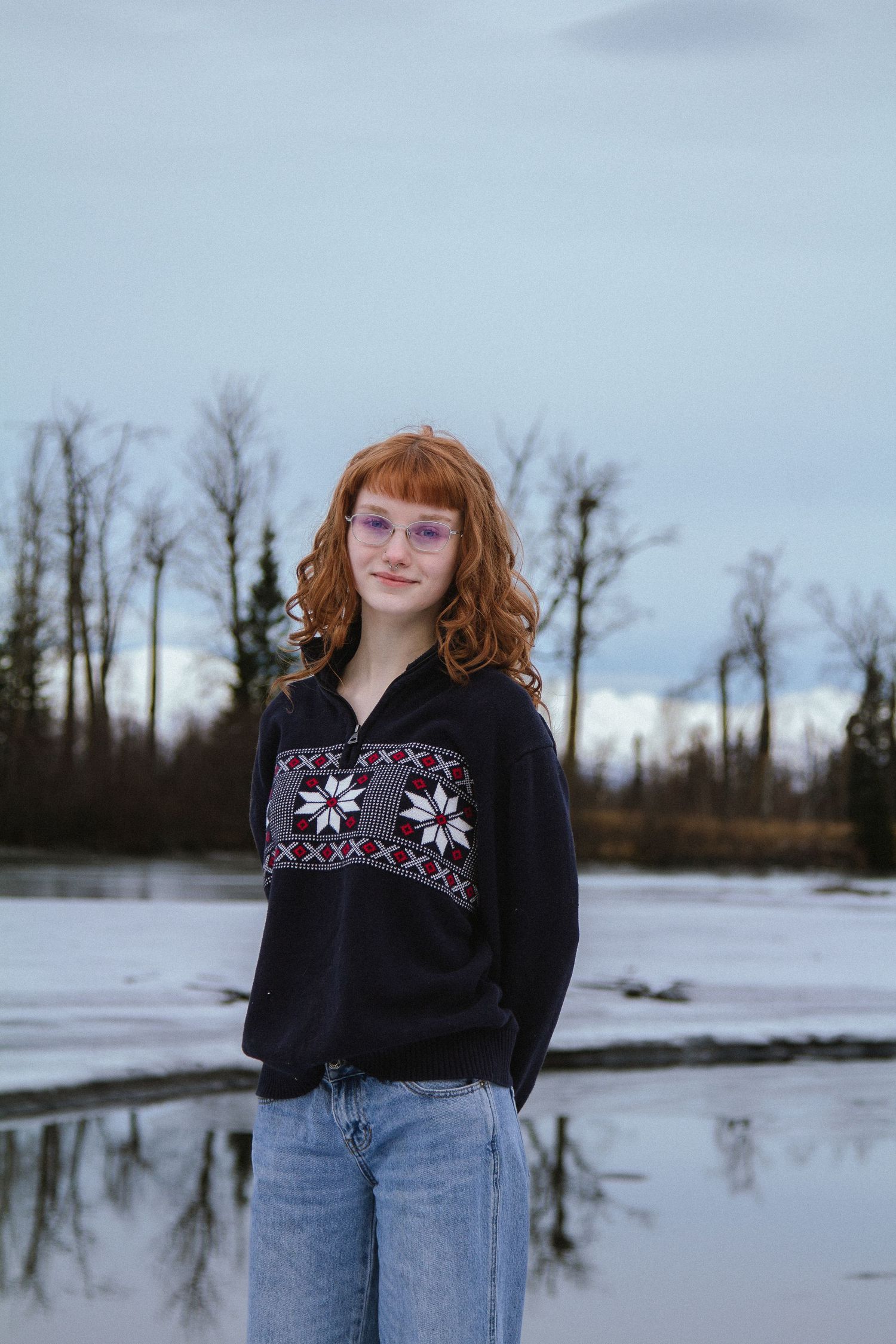 A person in a black sweater with floral embroidery and jeans poses outdoors against a snowy winter landscape.