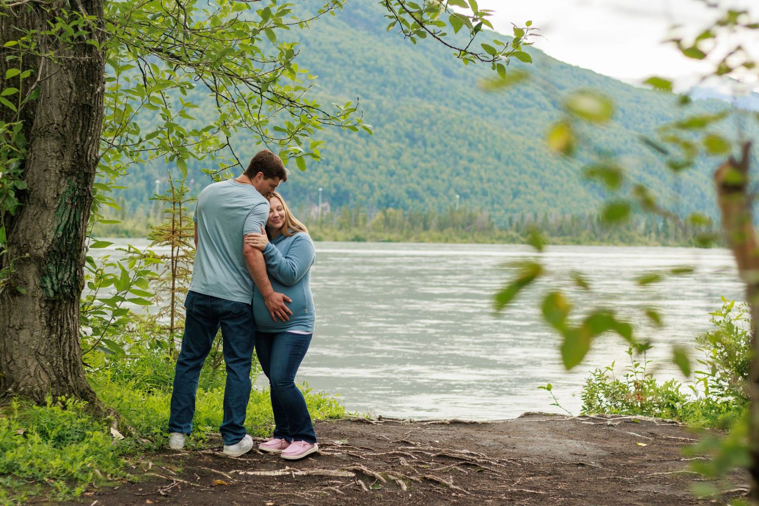 A couple shares a romantic moment along a scenic lakeside trail surrounded by lush green trees and mountains.