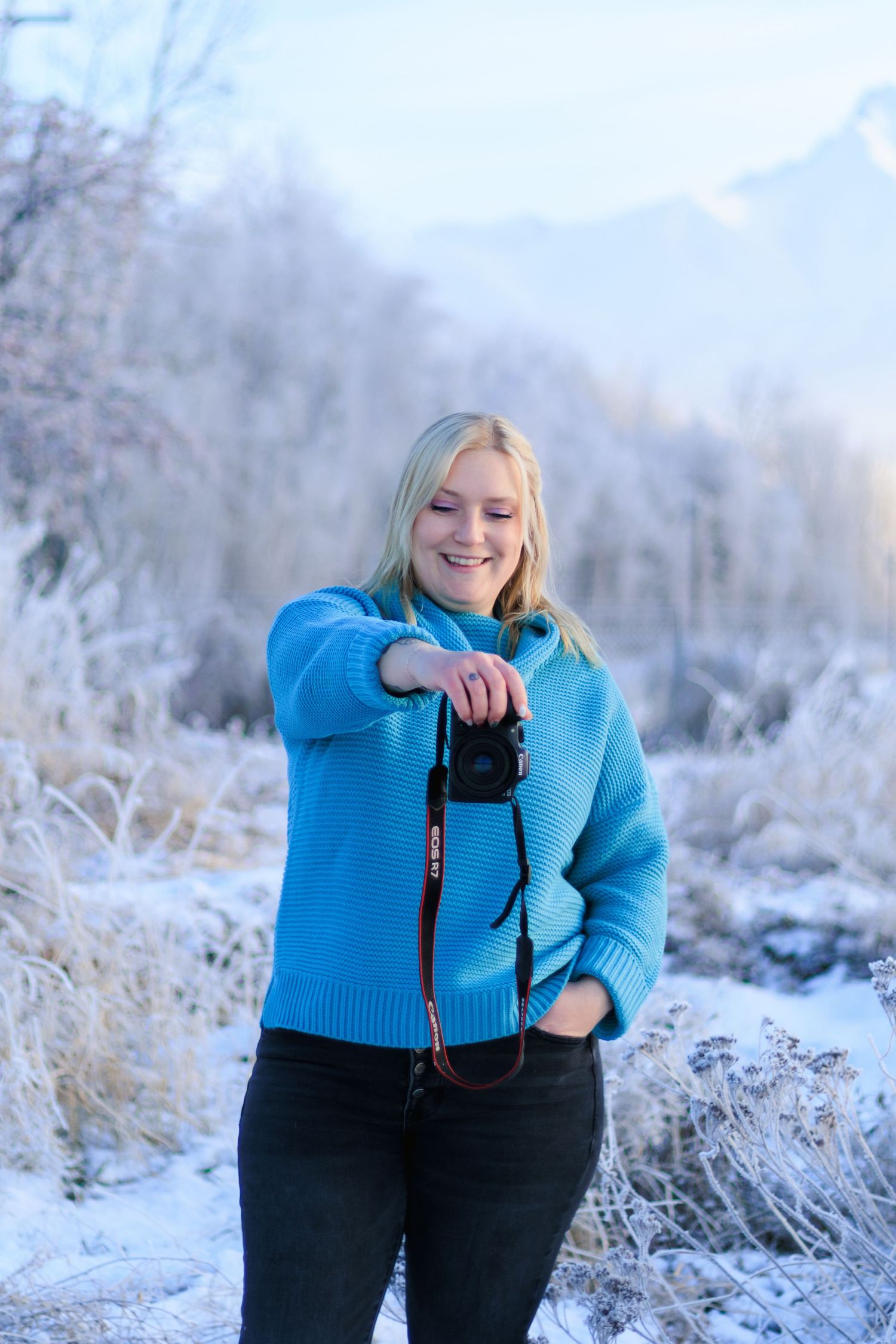 Person in bright blue sweater and black pants poses against snowy winter landscape with frosty vegetation.