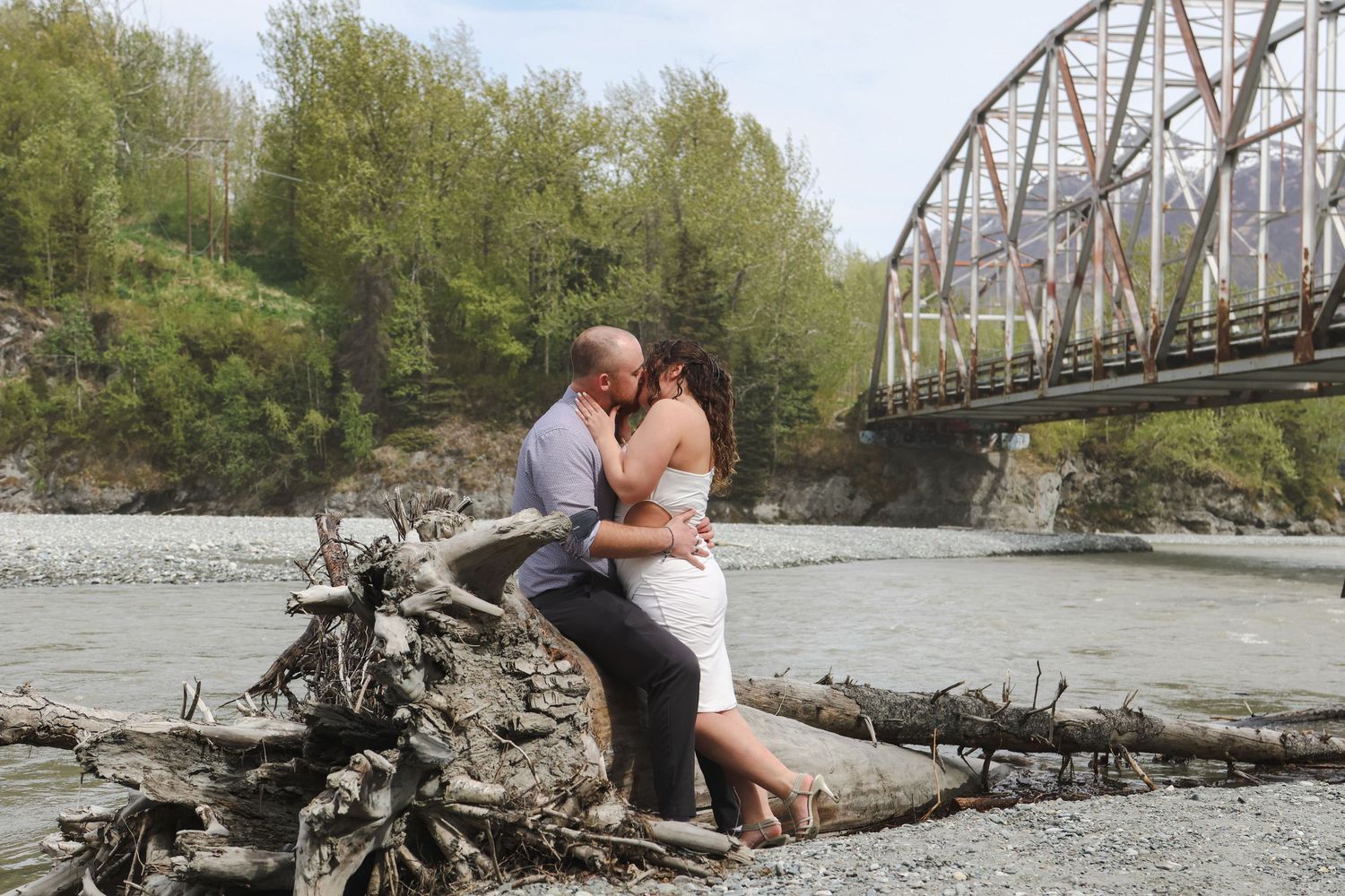 A romantic engagement photo session takes place by a rustic metal bridge over a river on a spring afternoon.