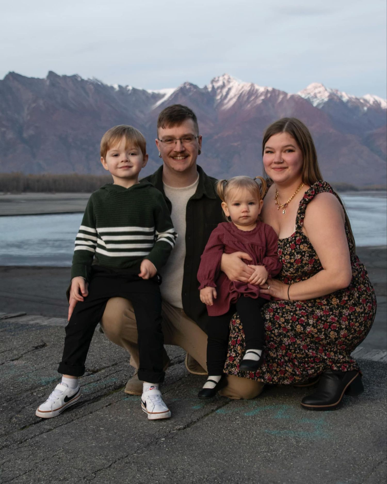 Family sitting together on a rocky beach with snow-capped mountains in the background at sunset.