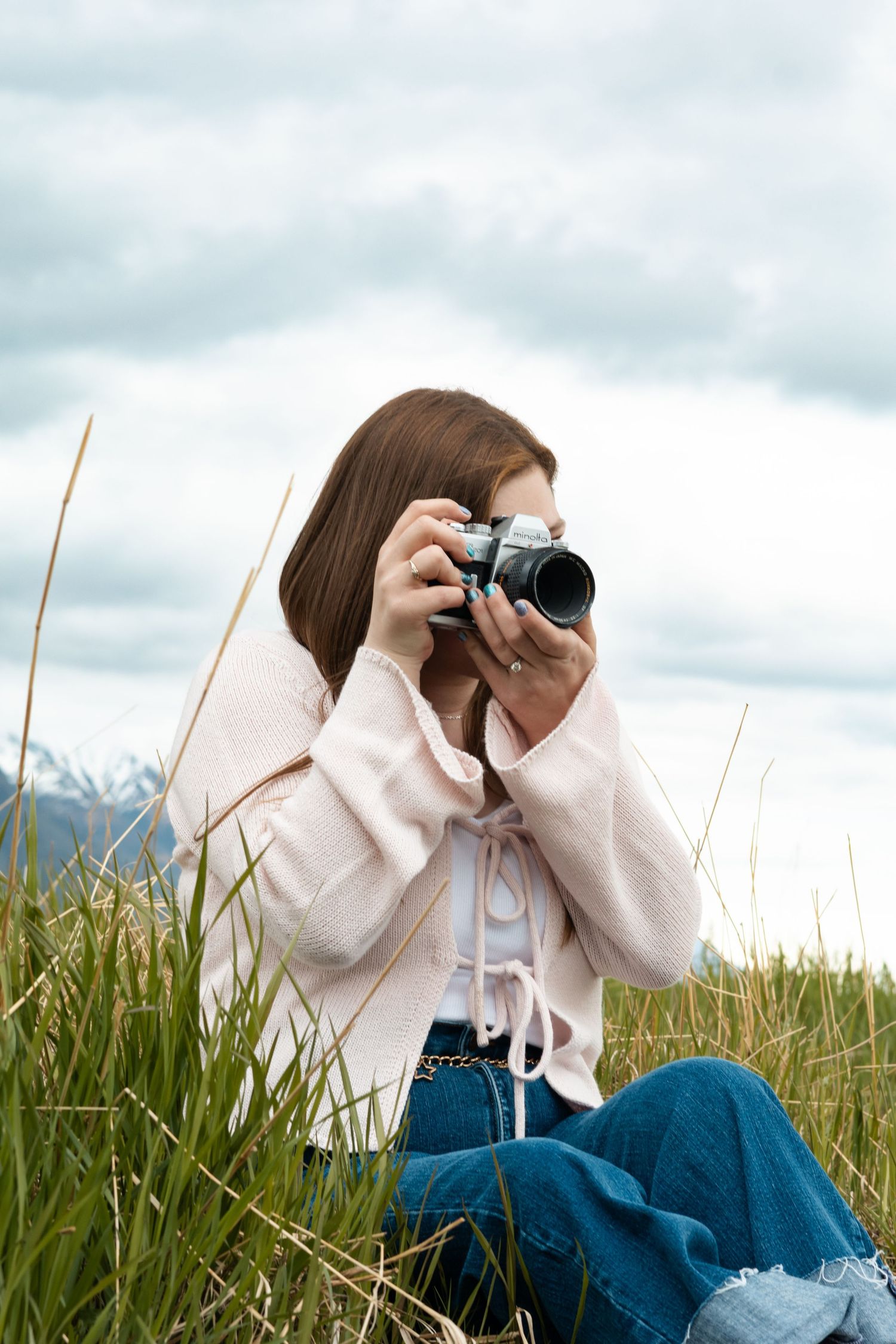 Young woman sitting in tall grass, holding a vintage film camera up to her eye, wearing a pink cardigan and jeans.