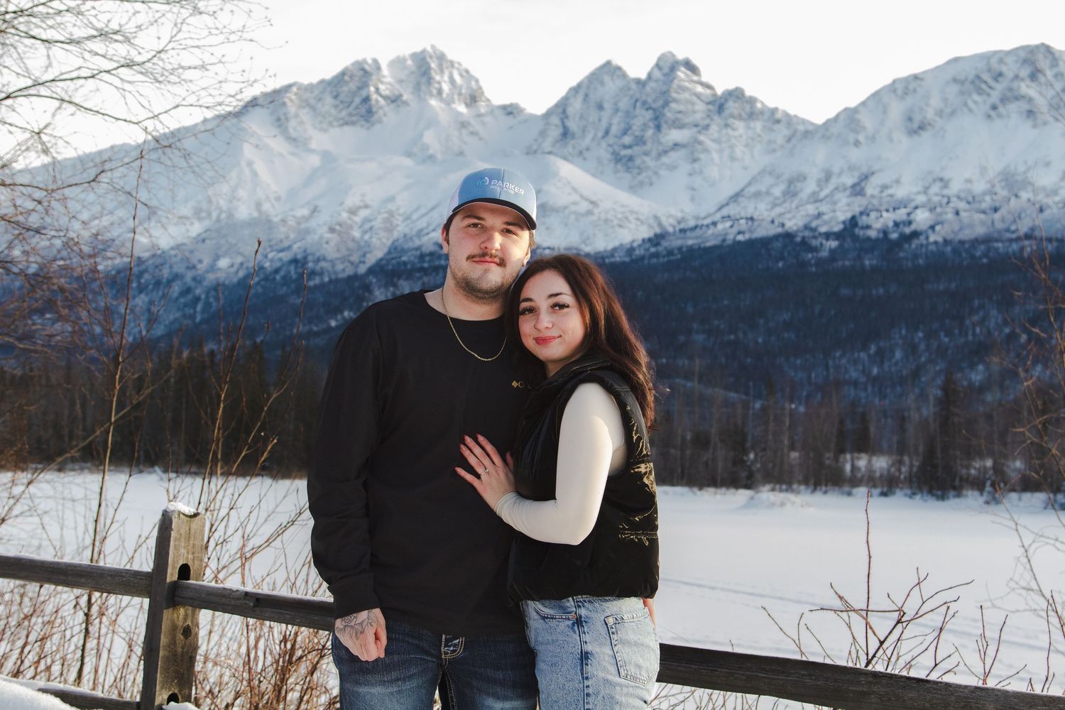 A couple embraces by a wooden fence with snow-capped mountains and a winter landscape in the background.