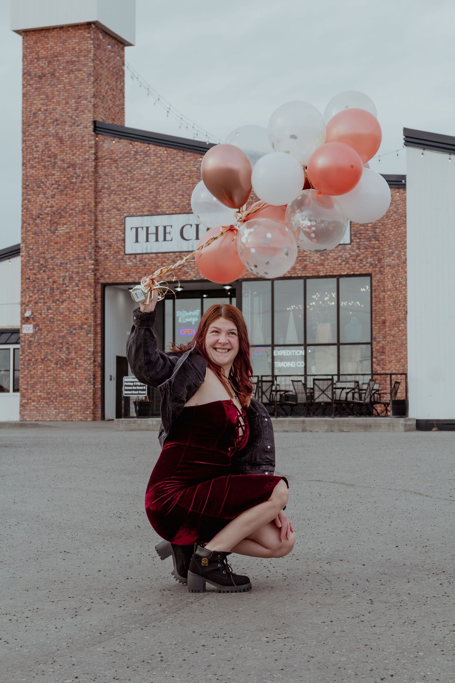 A person in a burgundy velvet dress poses playfully with pink and white balloons against a brick building exterior.