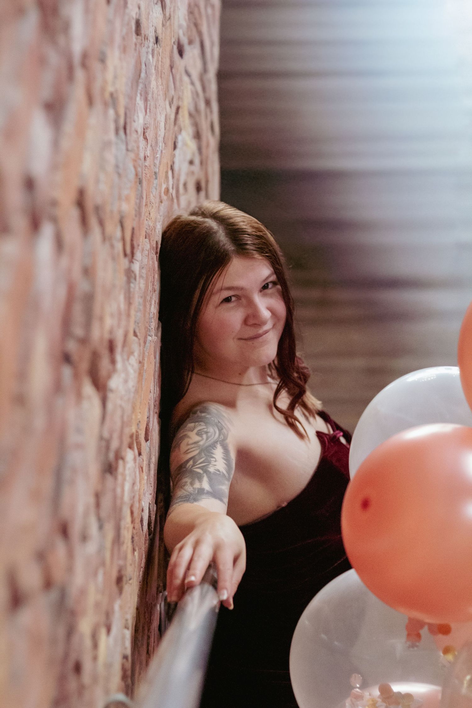 Portrait against brick wall with pink and white balloons featuring dark dress and visible arm tattoo.