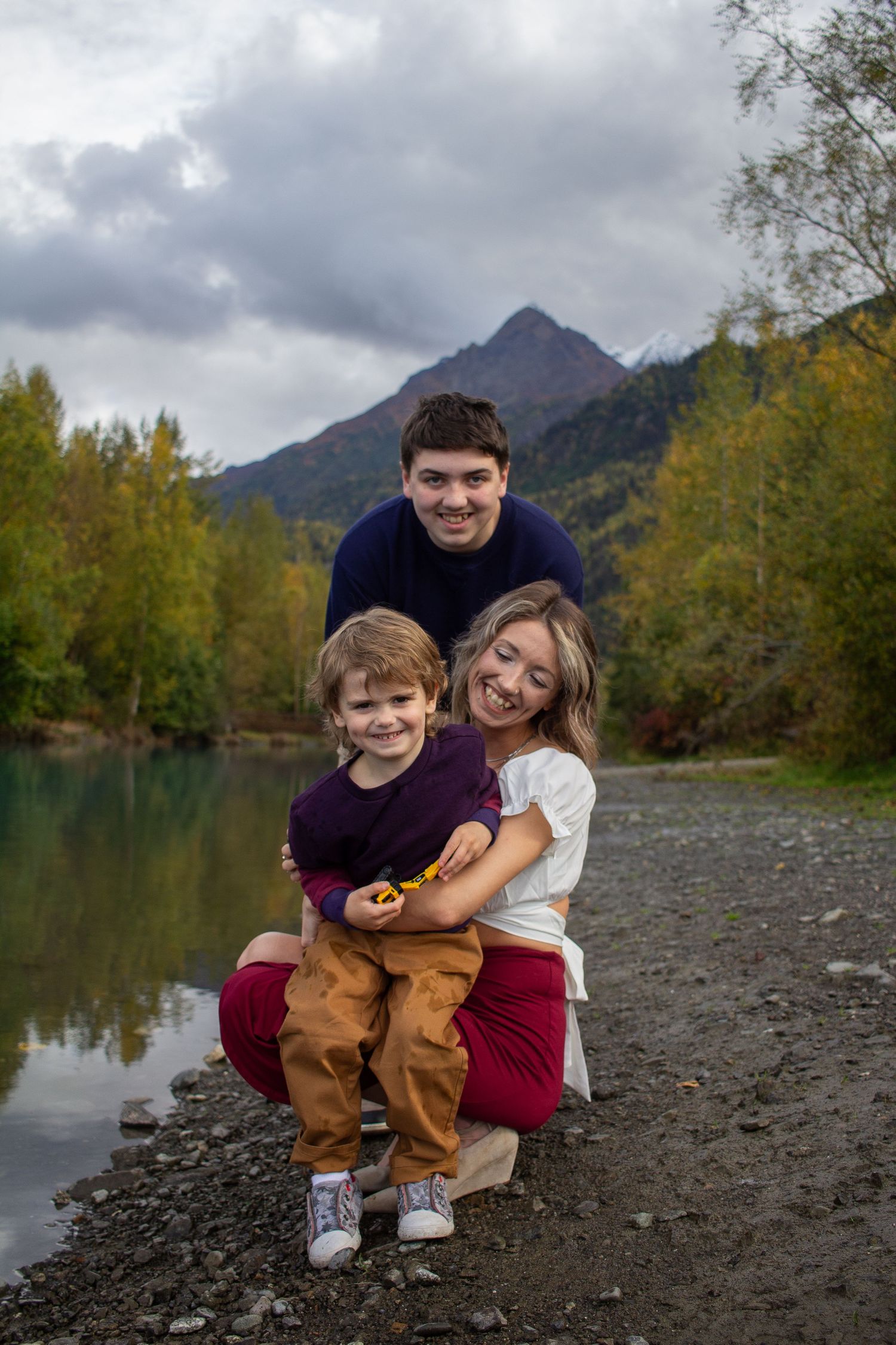 Two people sit together on a rocky shoreline with mountains and fall foliage reflected in the calm lake water.
