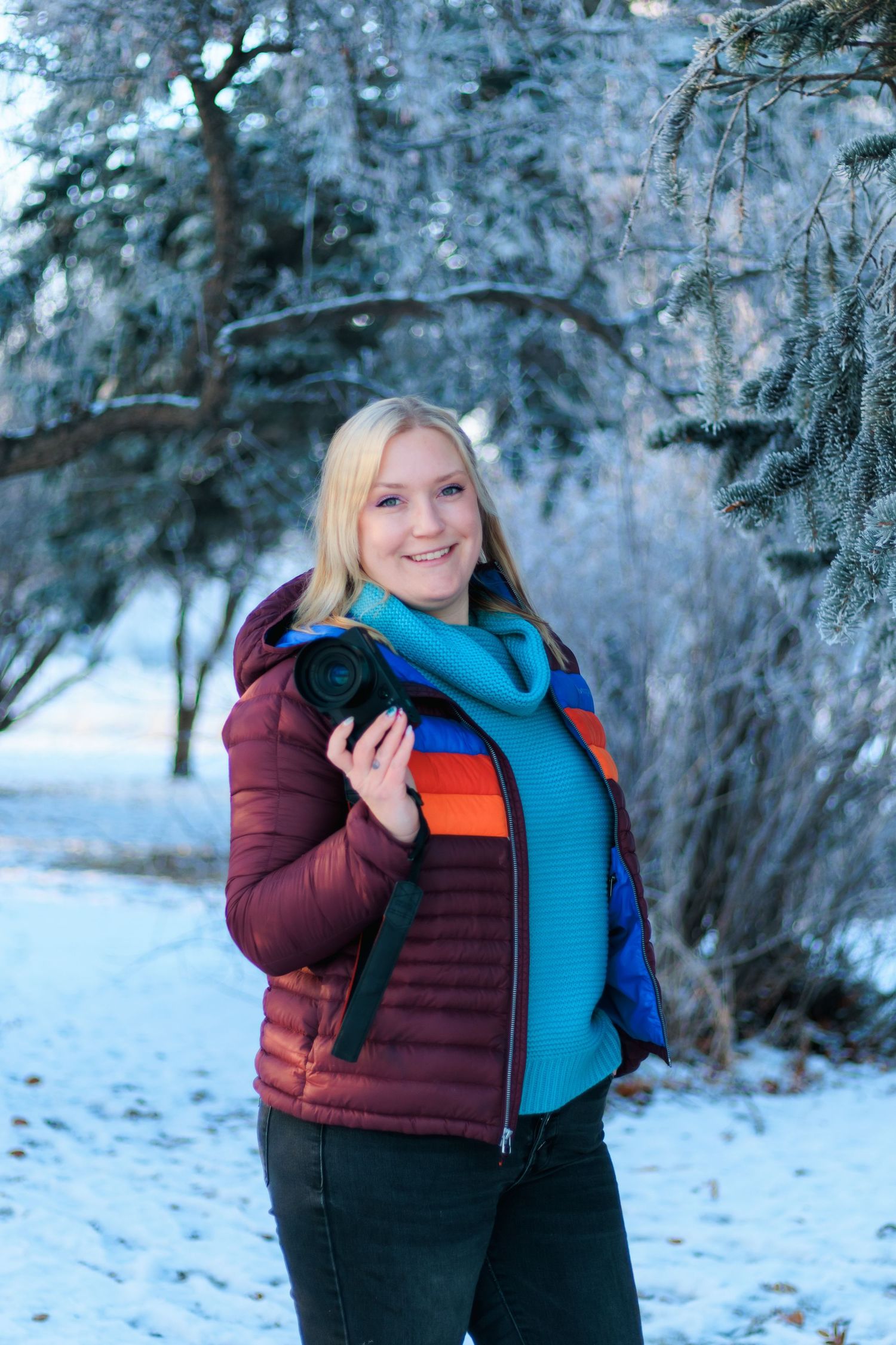 A person wearing a burgundy winter coat and bright blue scarf poses in a snowy winter landscape with evergreen trees.