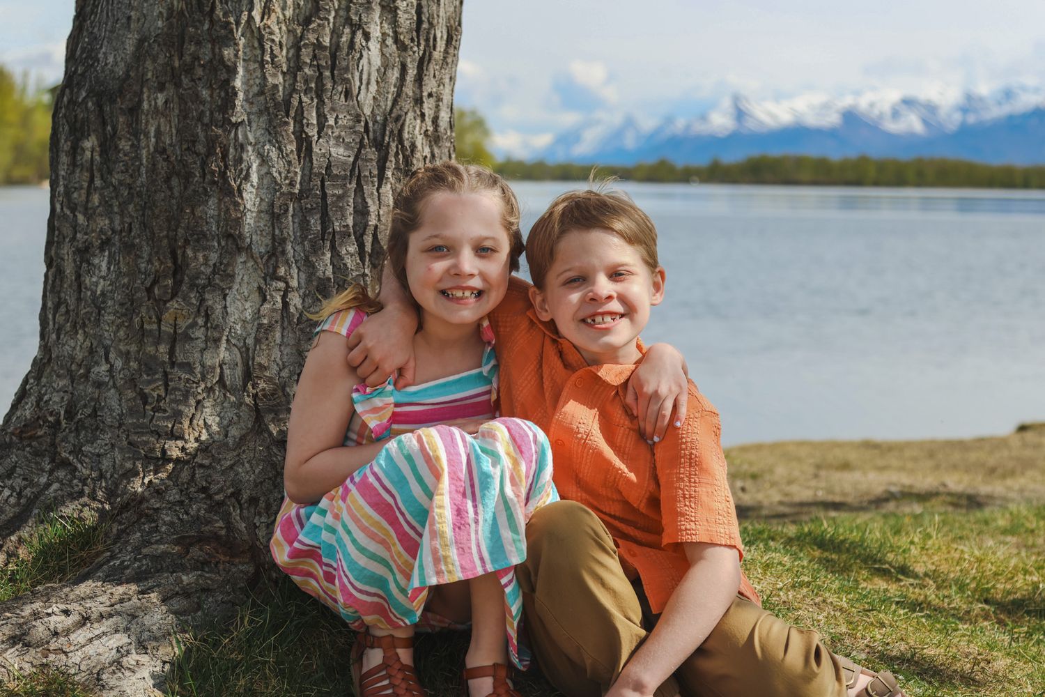 Two children sit together by a tree trunk near a lake with snow-capped mountains in the background.