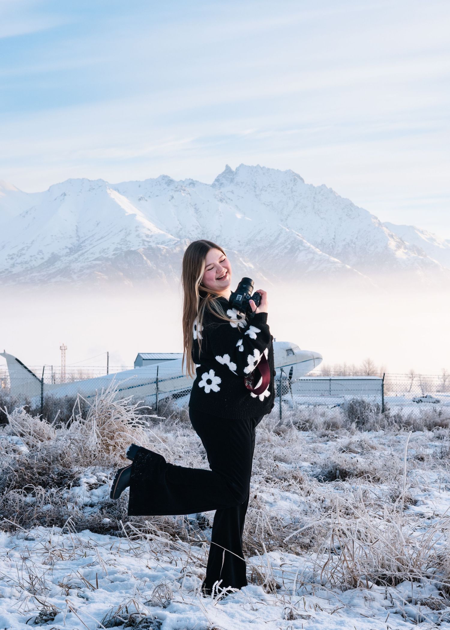 A winter landscape with snow-covered ground and mountains features someone in a black sweater posing playfully against the scenery.