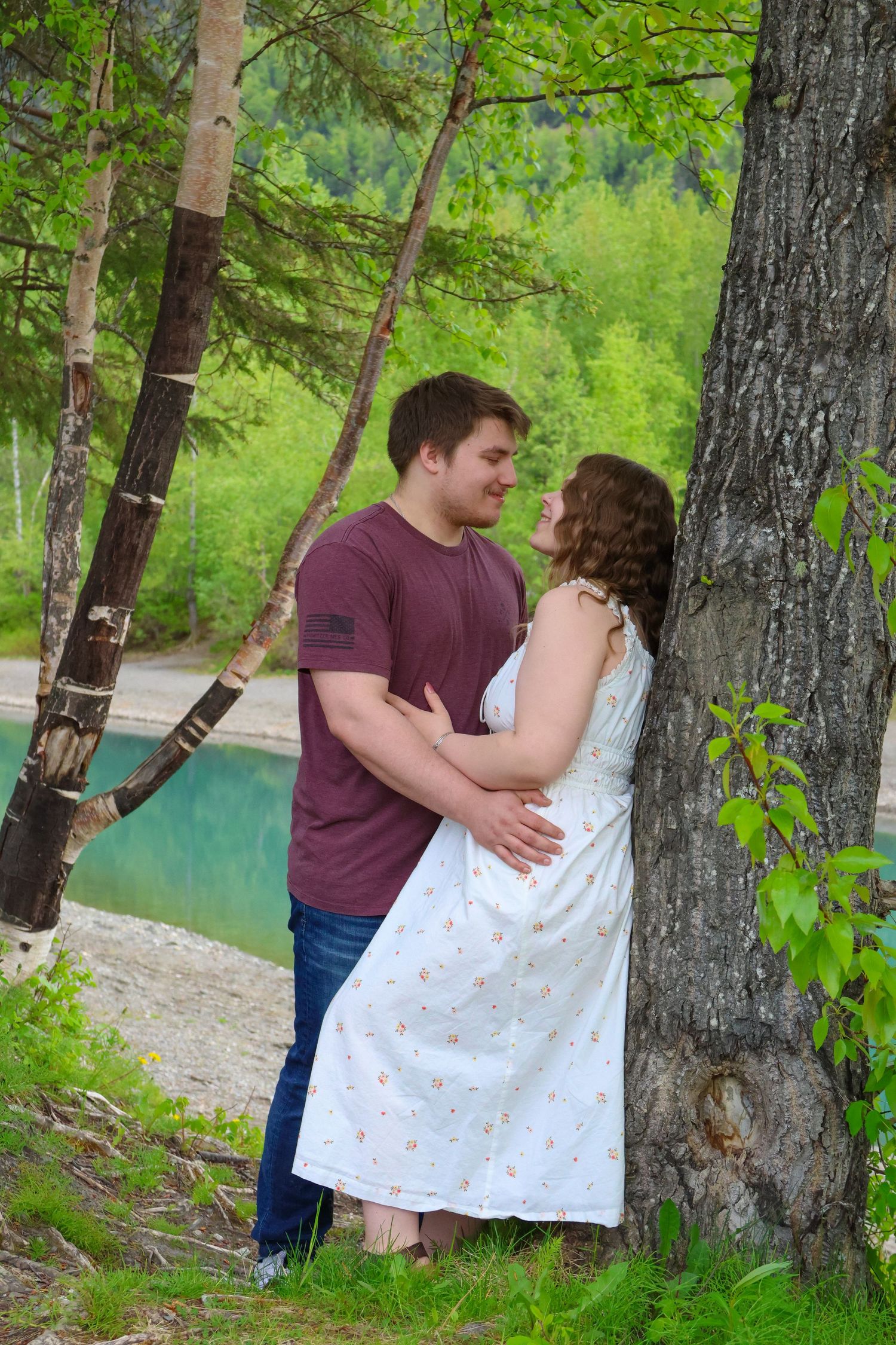 A romantic couple embraces by a tree near a turquoise lake surrounded by lush green forest.